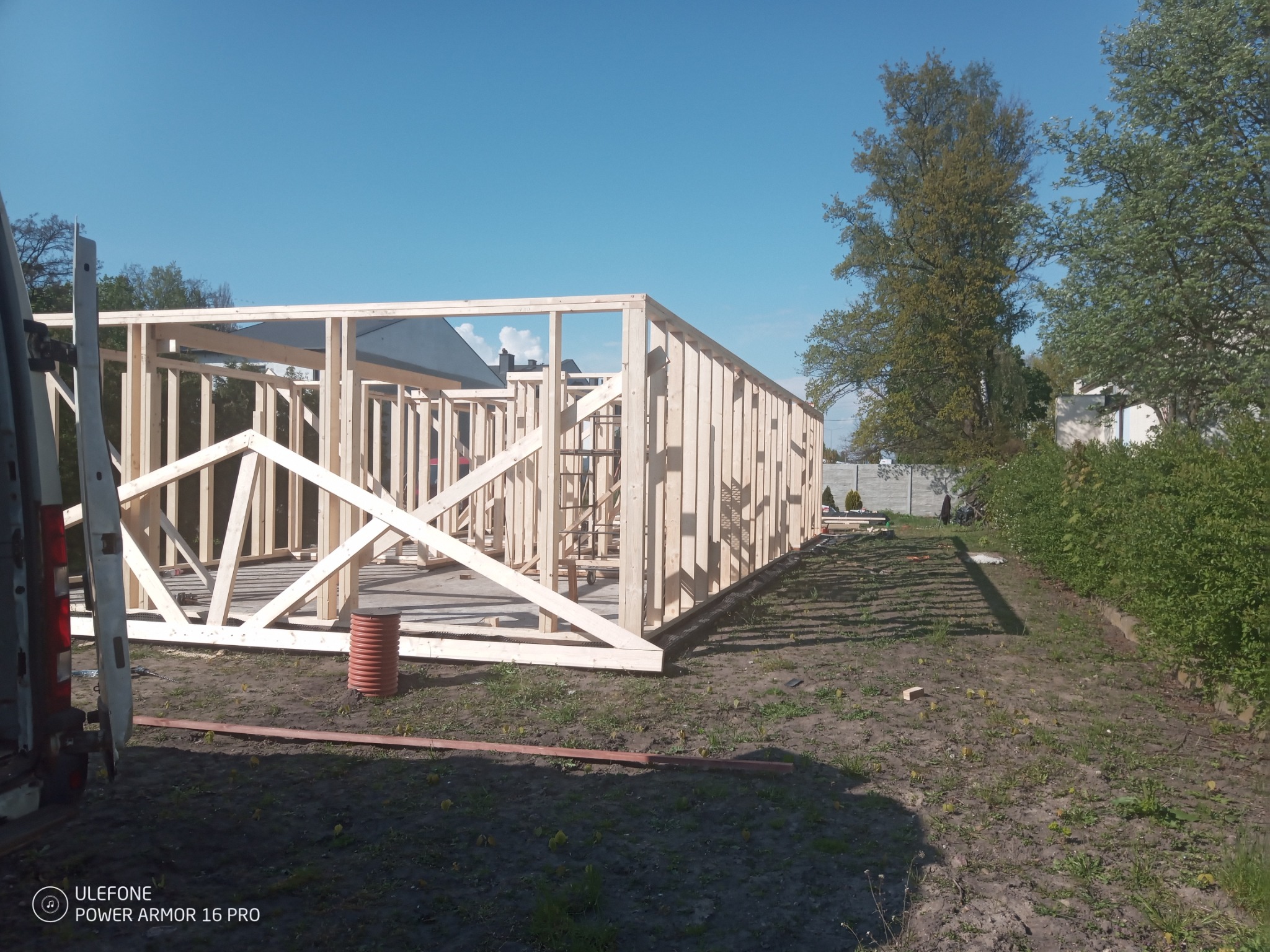 Drewniana konstrukcja szkieletowa domu w trakcie budowy, widoczny szkielet ścian i dachu na tle błękitnego nieba, z widocznym fragmentem samochodu dostawczego po lewej stronie.