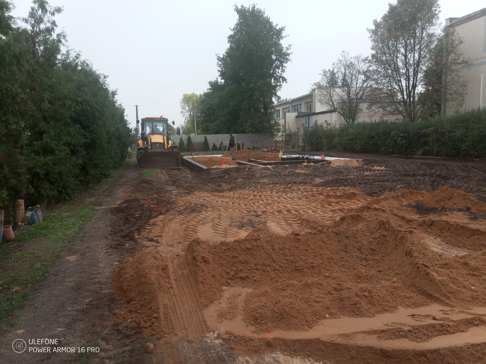 Żółta ładowarka czołowa na placu budowy, widoczne wykopane fundamenty i pracownik z łopatą, w tle budynek i drzewa.