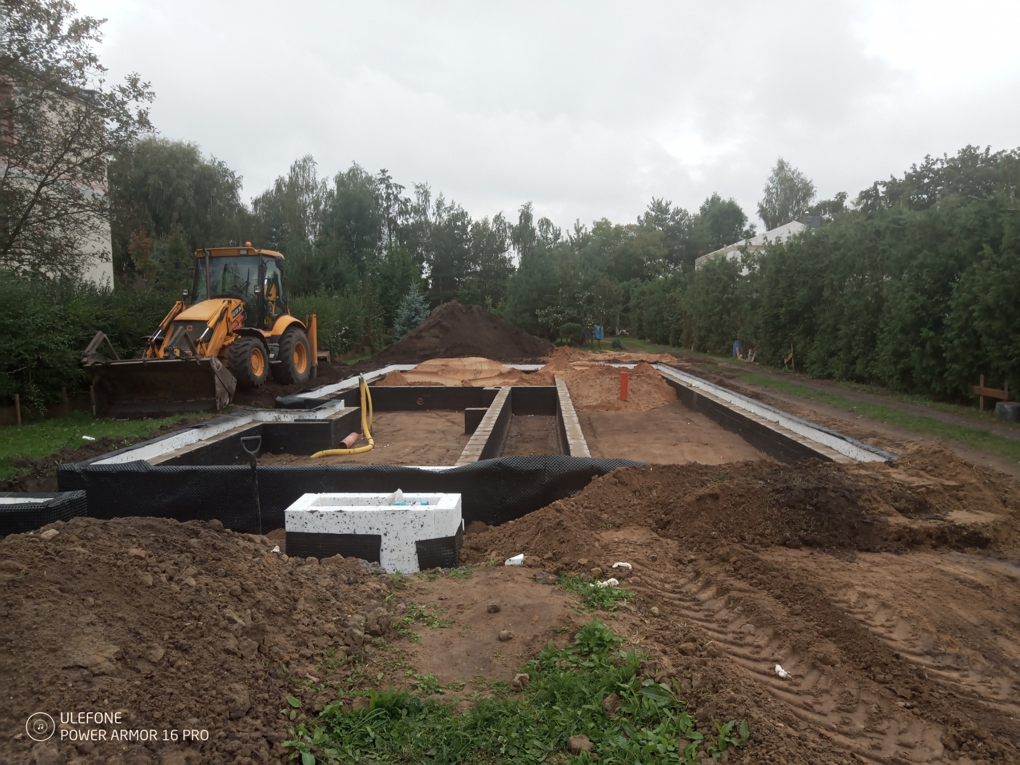Przygotowanie terenu pod budowę: wykopane fundamenty z izolacją, widoczny żółty traktor i hałdy ziemi na tle zieleni i pochmurnego nieba.
