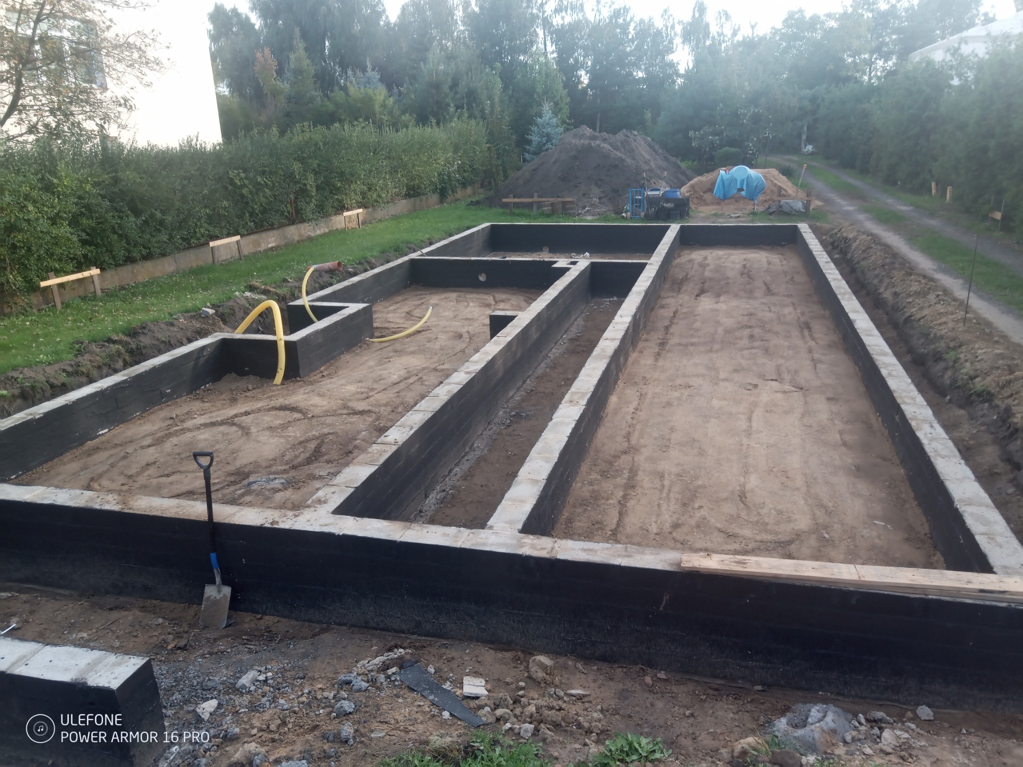 Zalane betonem fundamenty domu jednorodzinnego z hydroizolacją, widoczny wykop i pryzma ziemi w tle, szpadel wbity w ziemię.
