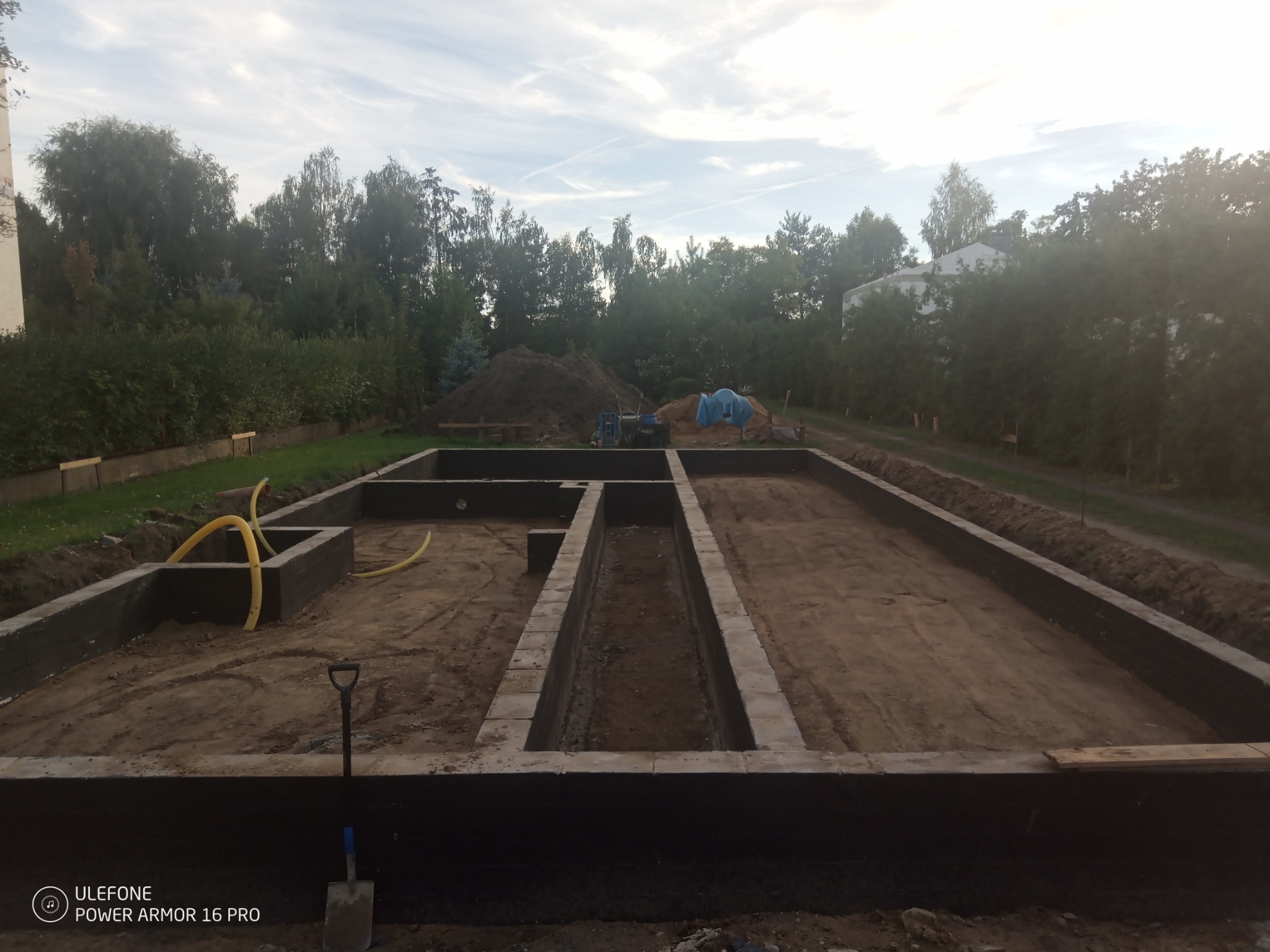 Zalane fundamenty domu jednorodzinnego z widocznymi żółtymi rurami instalacyjnymi i szpadlem w rogu, na tle zieleni i nieba z chmurami.