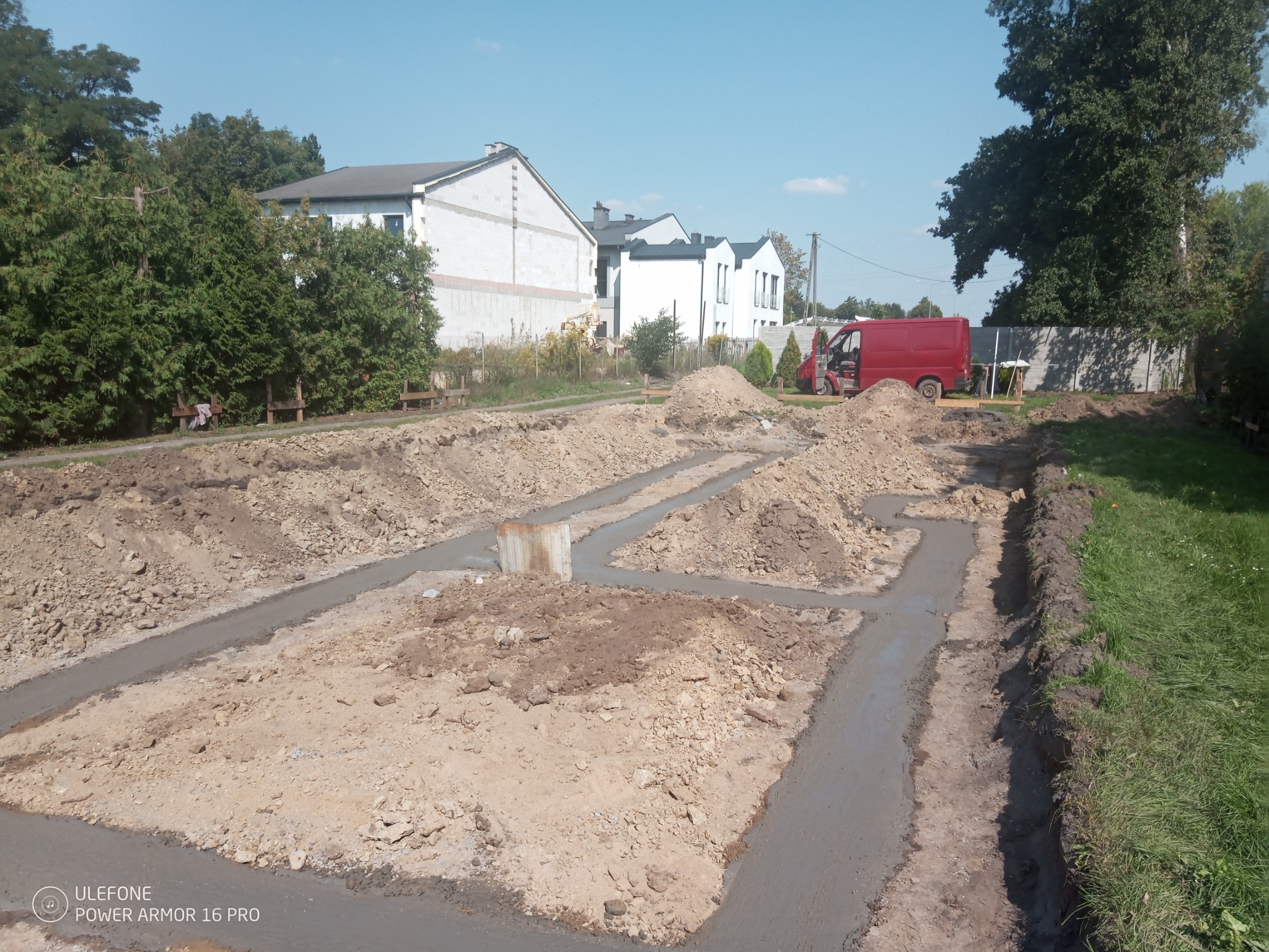 Zalane betonem wykopy pod fundamenty domu jednorodzinnego, widoczny fragment zbrojenia, w tle budynki mieszkalne i czerwony samochód dostawczy.