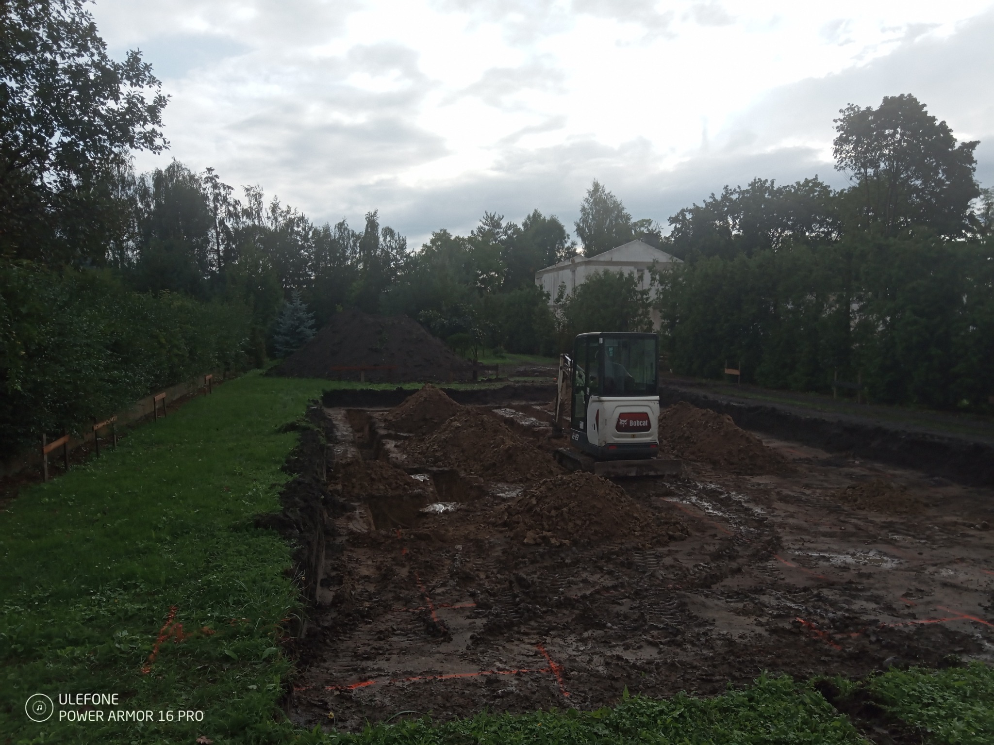 Mini koparka Bobcat na placu budowy, przygotowując teren pod fundamenty. Widoczne wykopane rowy i hałdy ziemi, w tle zieleń i budynek.