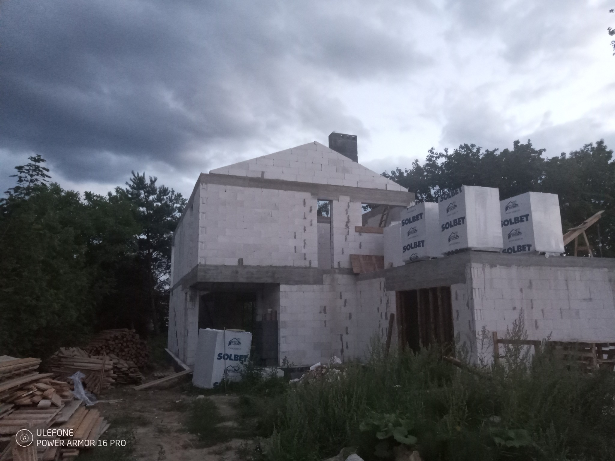 Surowy stan dwukondygnacyjnego domu z białych bloczków Solbet, widoczne sterty drewna i materiałów budowlanych, pochmurne niebo w tle.