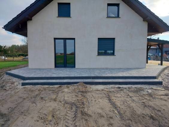 Nowo ułożony, ciemnoszary taras z kostki brukowej przed domem z szarą elewacją, widoczne dwa stopnie i przygotowany teren pod dalsze prace ziemne.