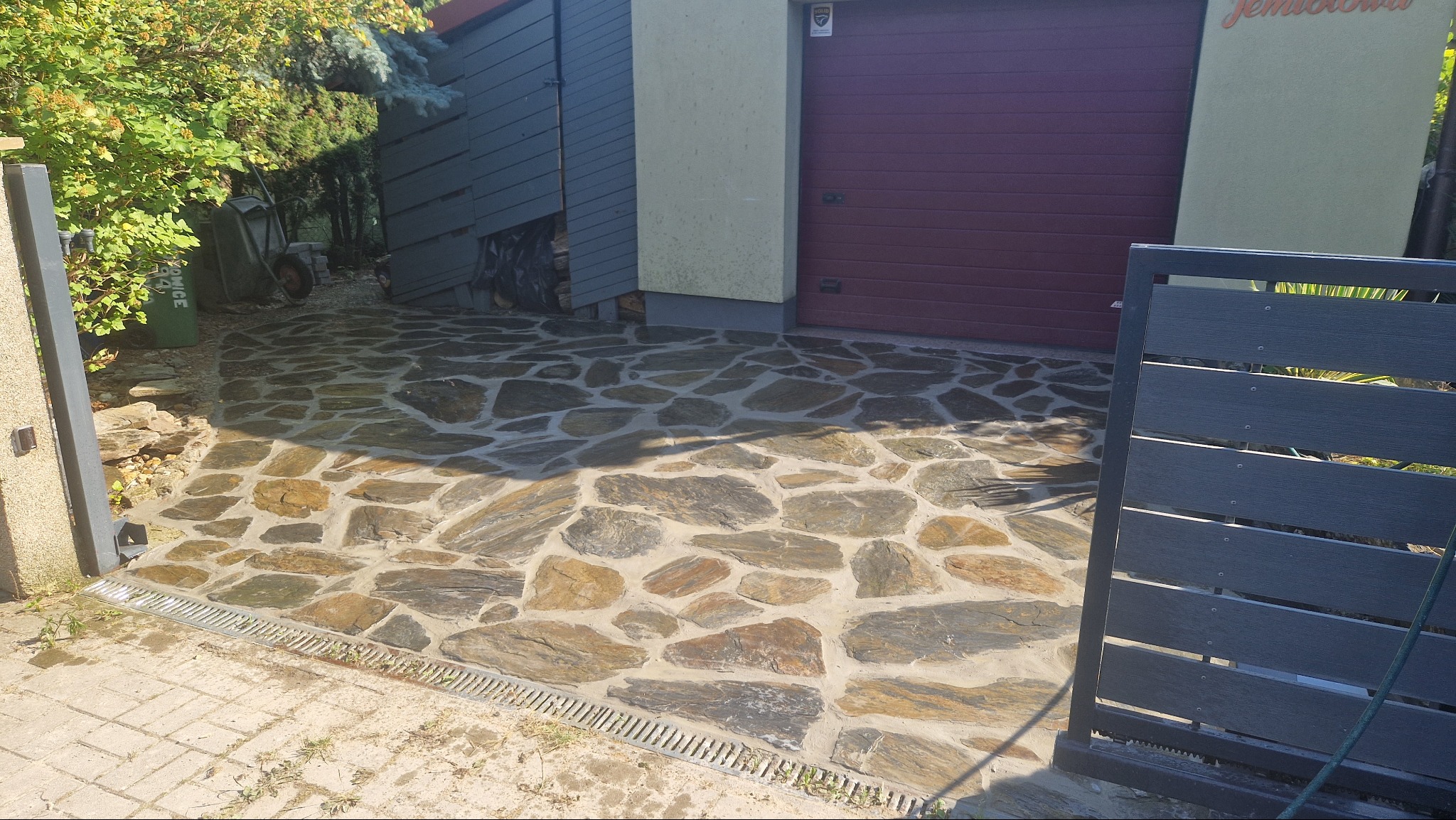 Nieregularna nawierzchnia z kamienia naturalnego w odcieniach szarości i brązu, ułożona przed garażem z bordową bramą, widoczna krata odpływowa przy bramie wjazdowej.