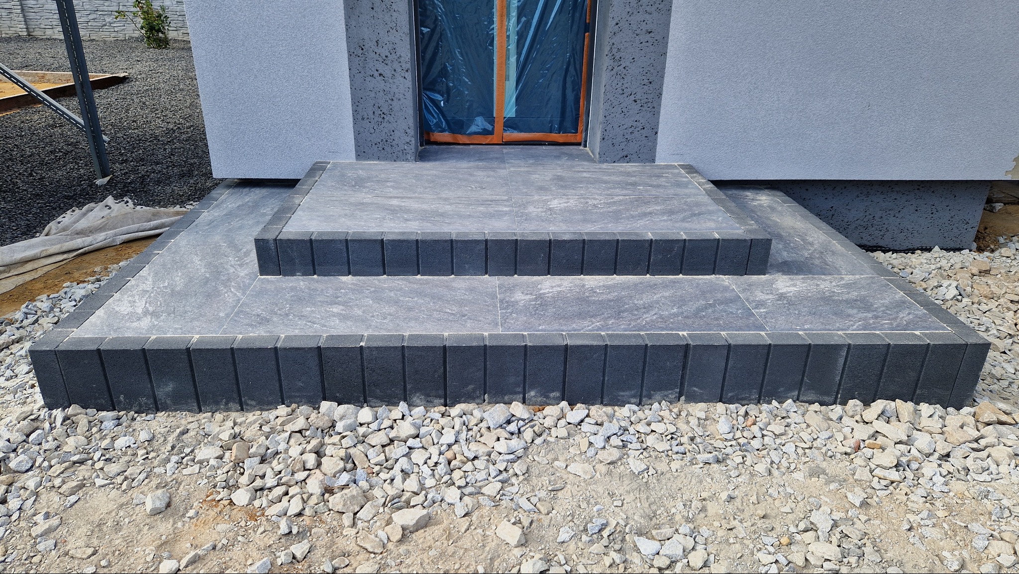 Nowe, dwustopniowe schody wejściowe wyłożone szarą kostką brukową, obrzeża z ciemnej kostki, widok z dołu na tle elewacji domu w trakcie wykończenia.