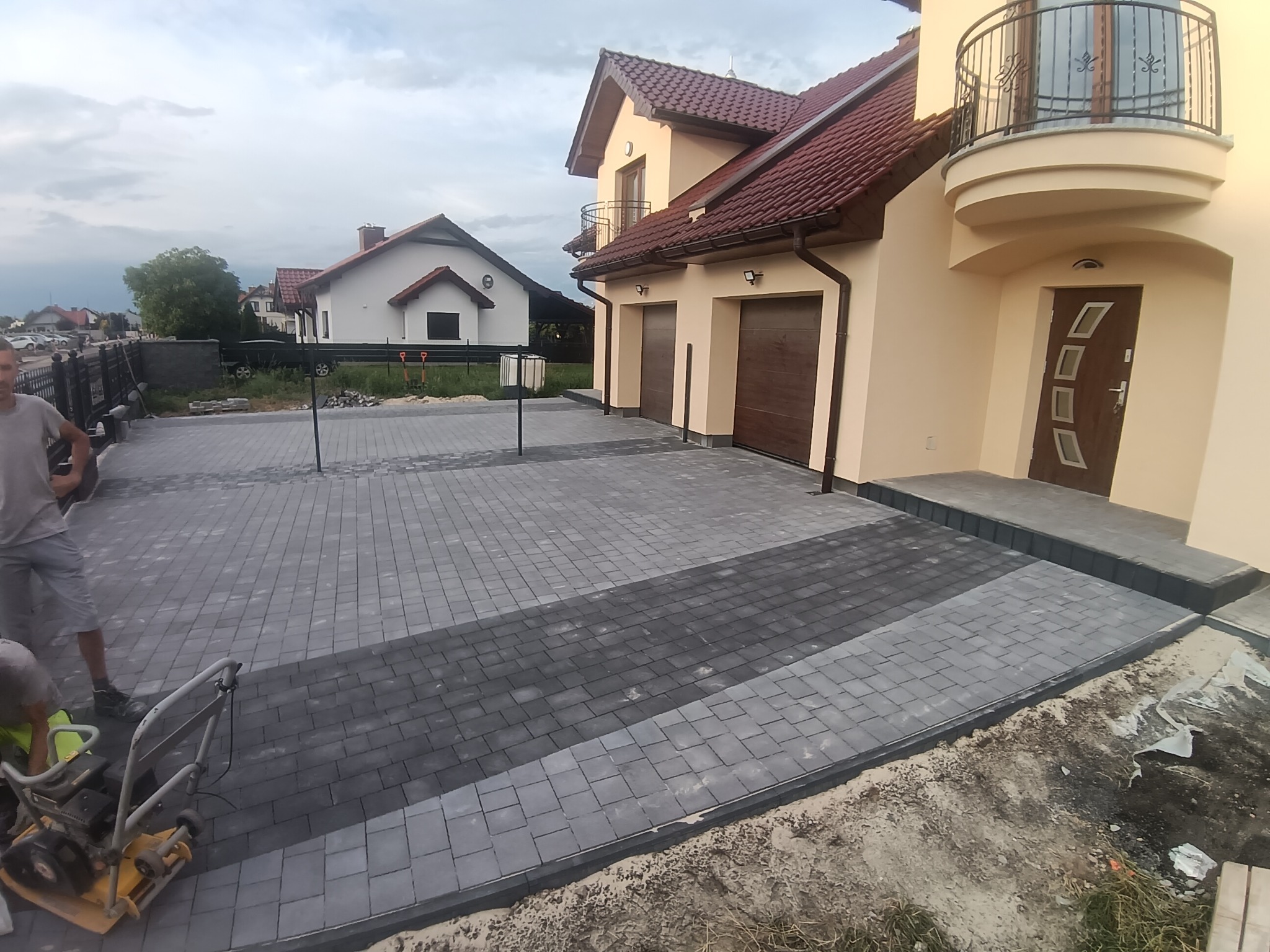 Ułożona kostka brukowa w dwóch odcieniach szarości tworząca wzór na podjeździe. W tle dom z beżową elewacją i brązowymi bramami garażowymi.