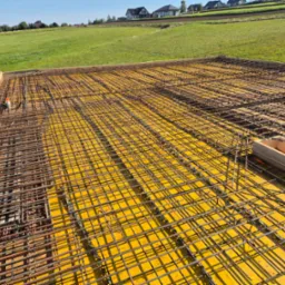 Zbrojenie stropu na żółtej płycie szalunkowej, widok z góry na tle zielonego pola i domów jednorodzinnych.