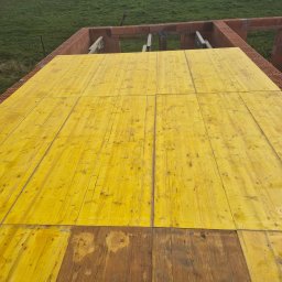 DOM-BUD MACIEJ SMALEC - Żółte deski szalunkowe tworzące strop nad parterem w trakcie budowy domu z czerwonej cegły, widok z góry na zielone pole w tle.