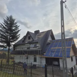 Wznoszenie dwukondygnacyjnego domu z garażem w bryle, widoczne rusztowania, częściowo położony dach z czarnej dachówki, druga część dachu w trakcie budowy z widoczną niebieską membraną,...