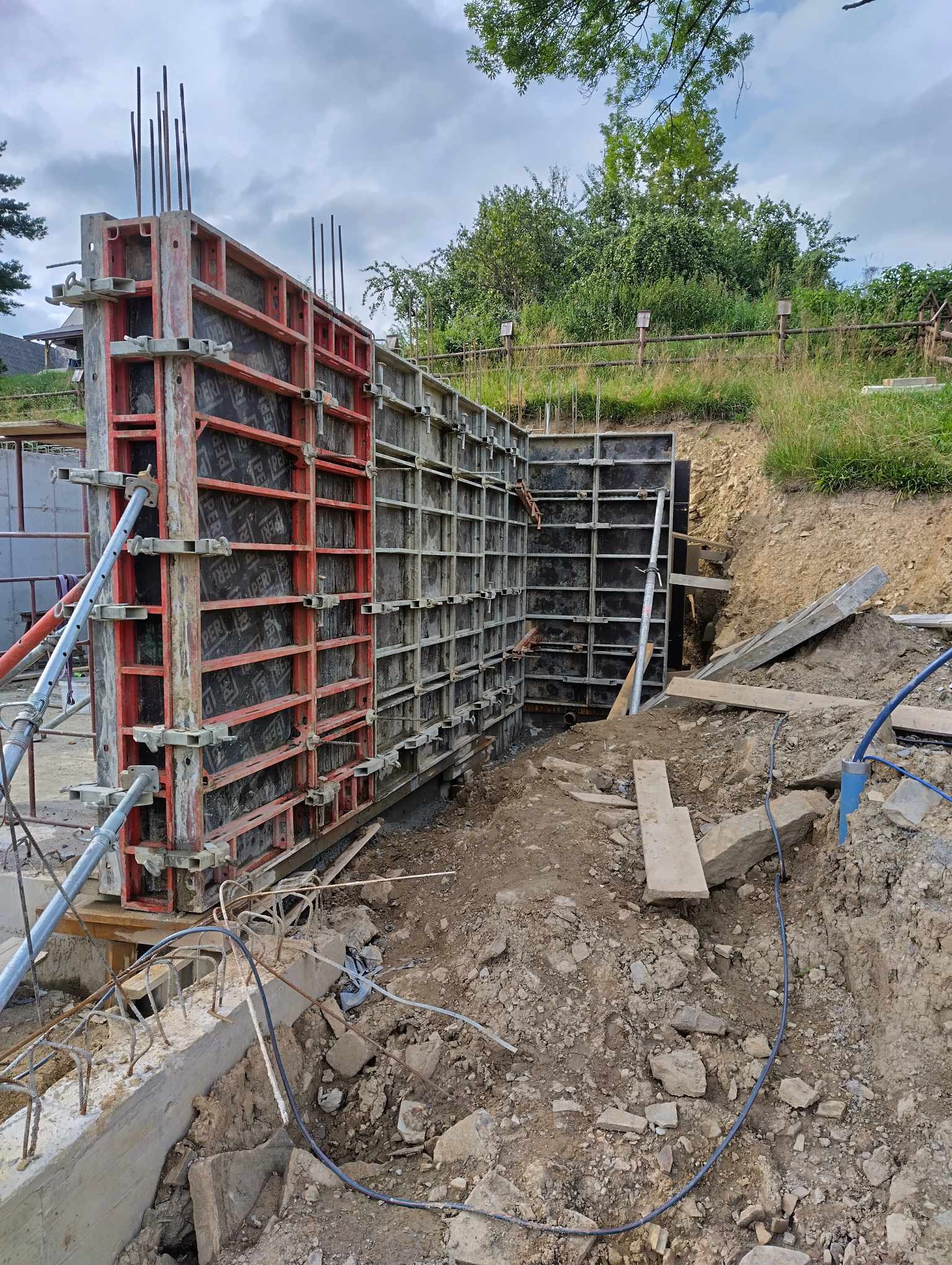 Szare i czerwone szalunki pionowe z widocznymi zbrojeniami, przygotowane do wylania betonu na fundamencie budynku w wykopie ziemnym.