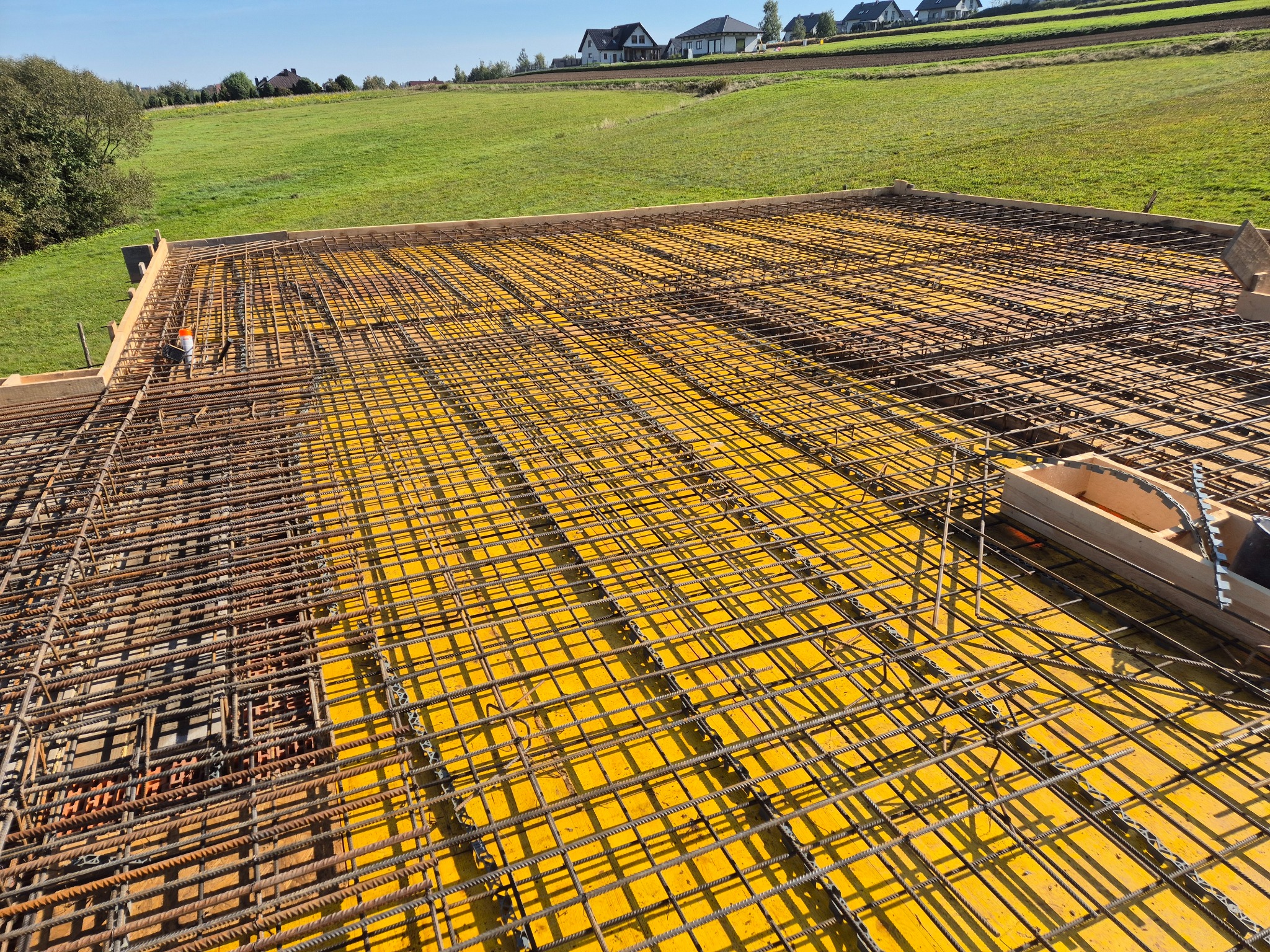 Zbrojenie stropu na żółtej płycie szalunkowej, widok z góry na tle zielonego pola i domów jednorodzinnych.
