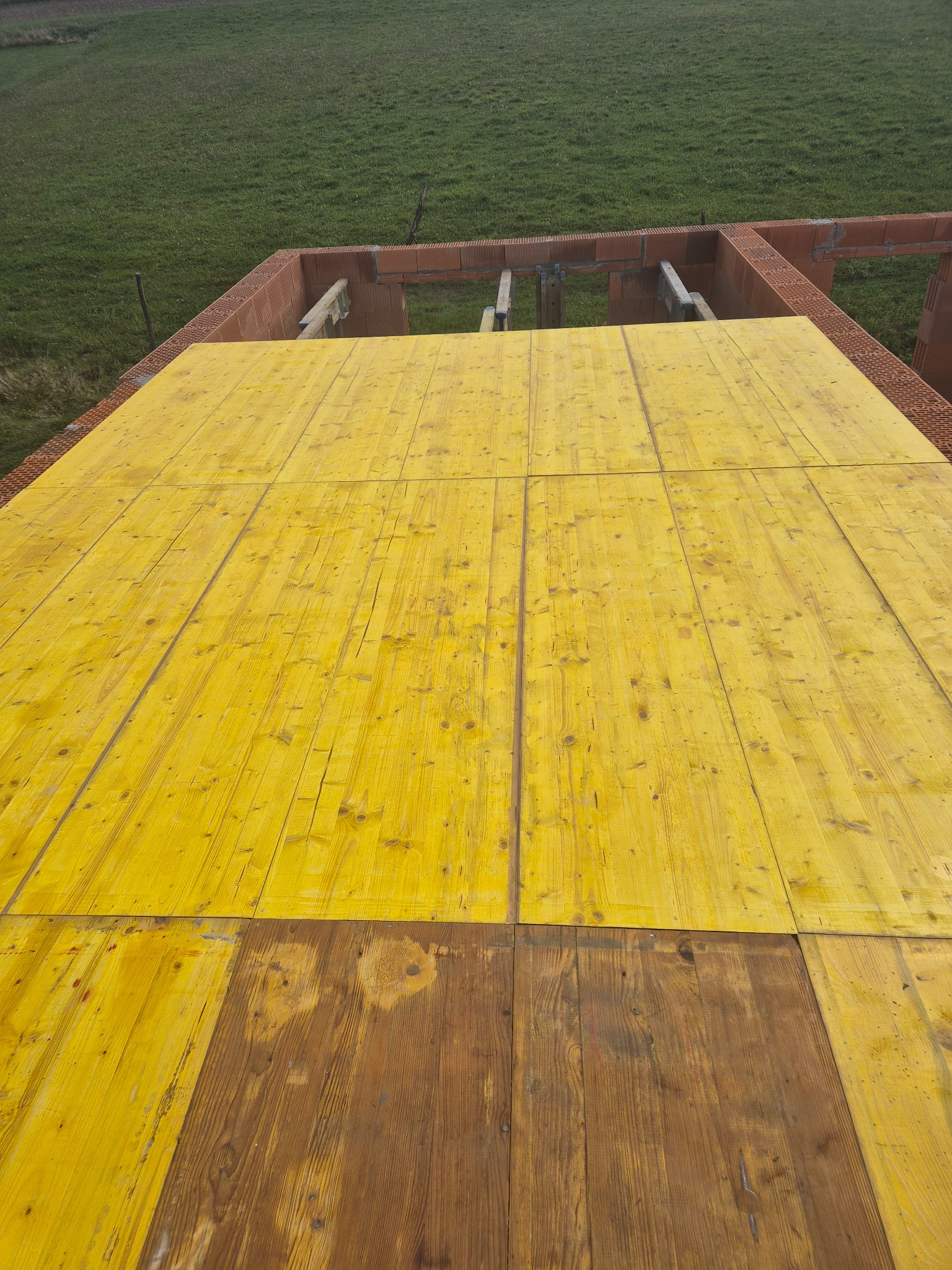 Żółte deski szalunkowe tworzące strop nad parterem w trakcie budowy domu z czerwonej cegły, widok z góry na zielone pole w tle.