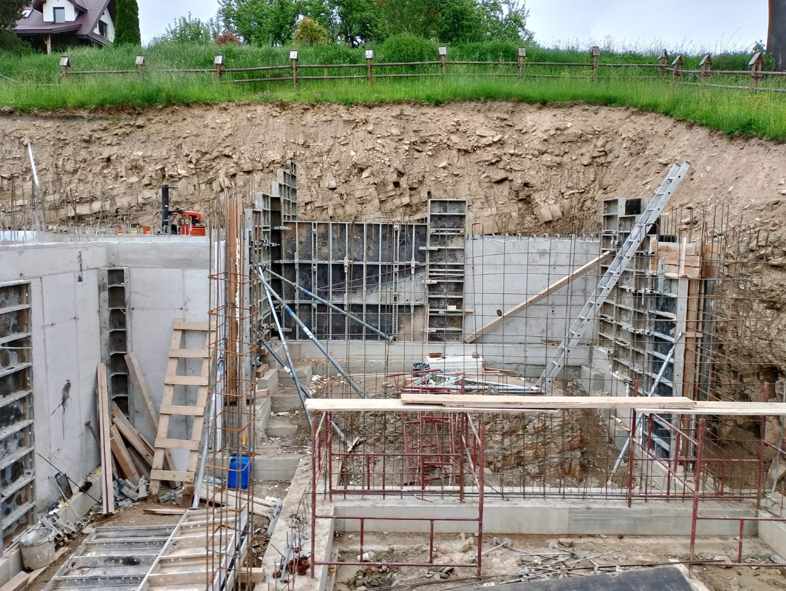 Wykop pod fundamenty z widocznymi szalunkami, zbrojeniami i częściowo wylanym betonem, na tle ziemi i zieleni