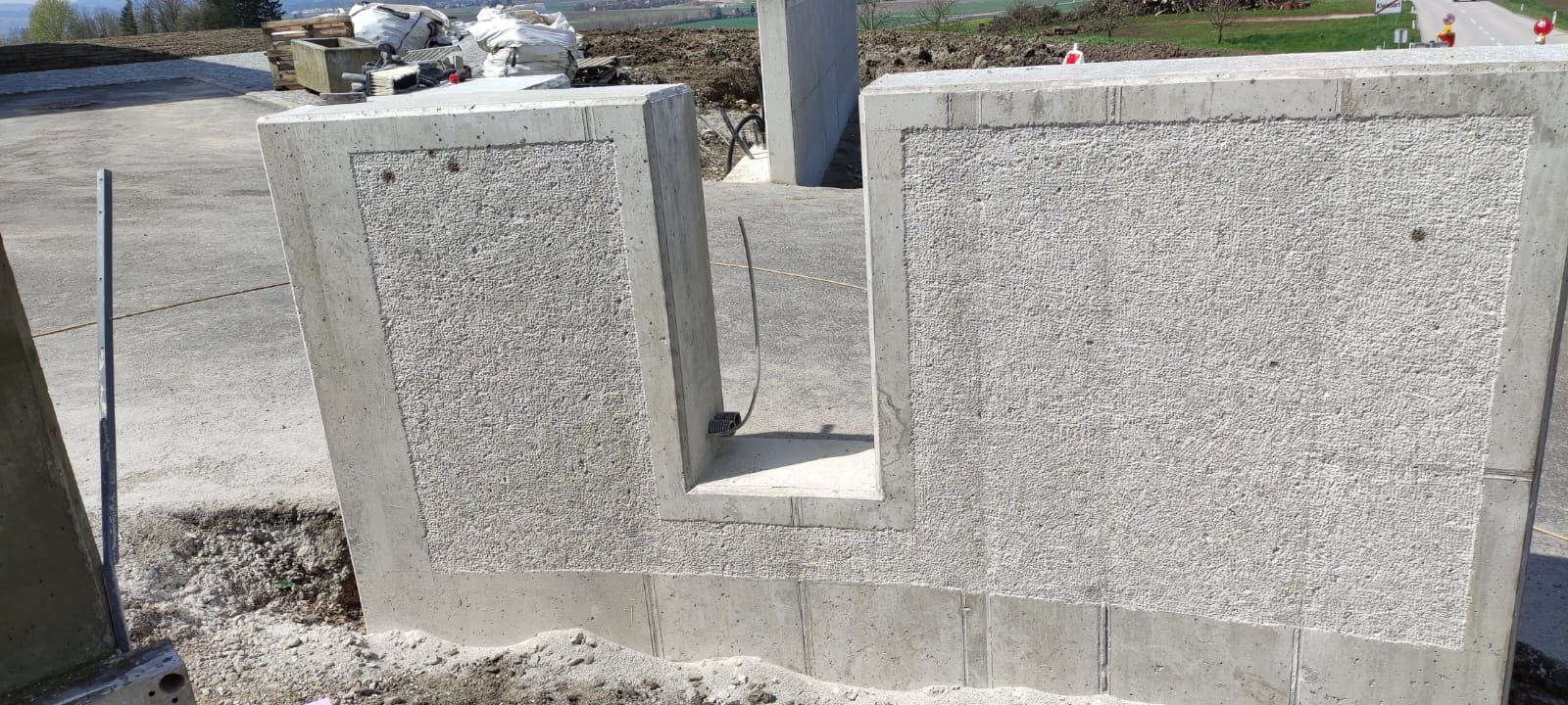 Surowy betonowy fundament z charakterystyczną fakturą i otworem na instalacje, widoczne ślady szalunku, tło z rozległym krajobrazem.