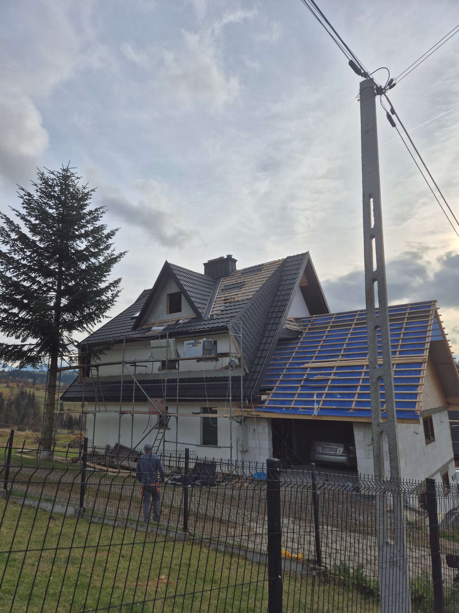 Wznoszenie dwukondygnacyjnego domu z garażem w bryle, widoczne rusztowania, częściowo położony dach z czarnej dachówki, druga część dachu w trakcie budowy z widoczną niebieską membraną,...