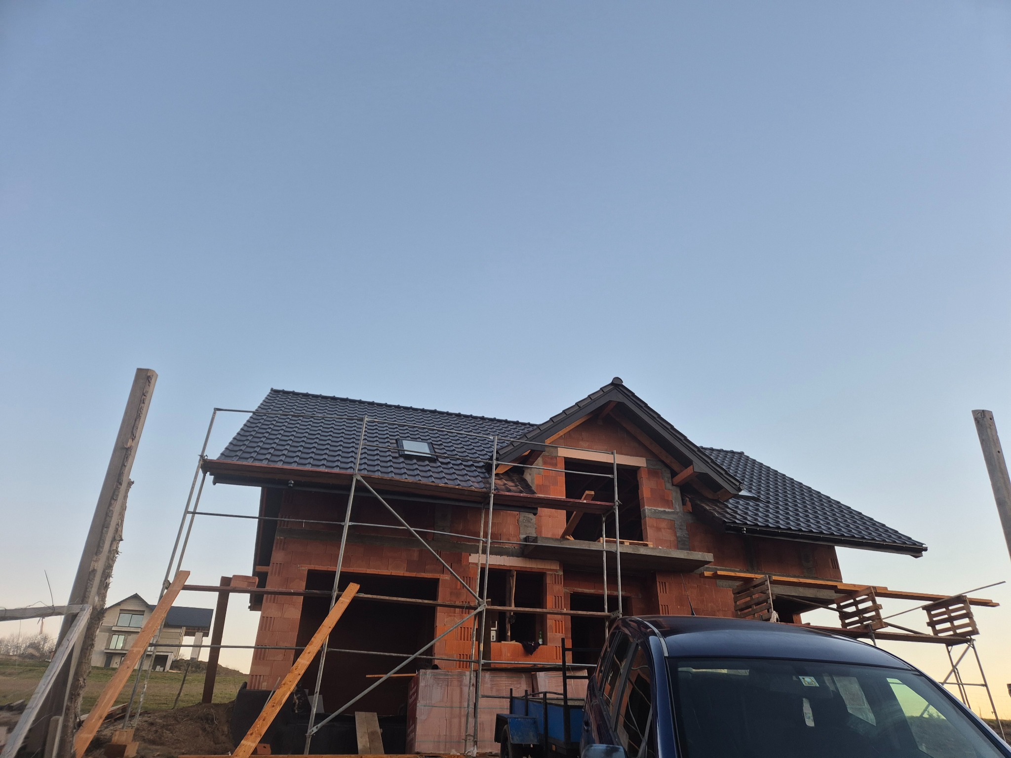 Wznoszony dwukondygnacyjny budynek z cegły ceramicznej, z ciemną dachówką, otoczony rusztowaniem, na tle błękitnego nieba, z fragmentem samochodu na pierwszym planie.