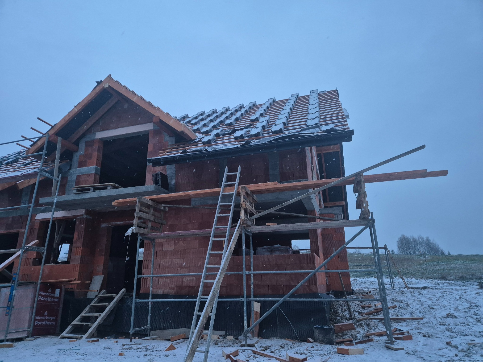 Niedokończony budynek z czerwonej cegły w zimowej scenerii, częściowo pokryty dachówką, z rusztowaniami i drabinką, widoczne bloczki Porotherm.