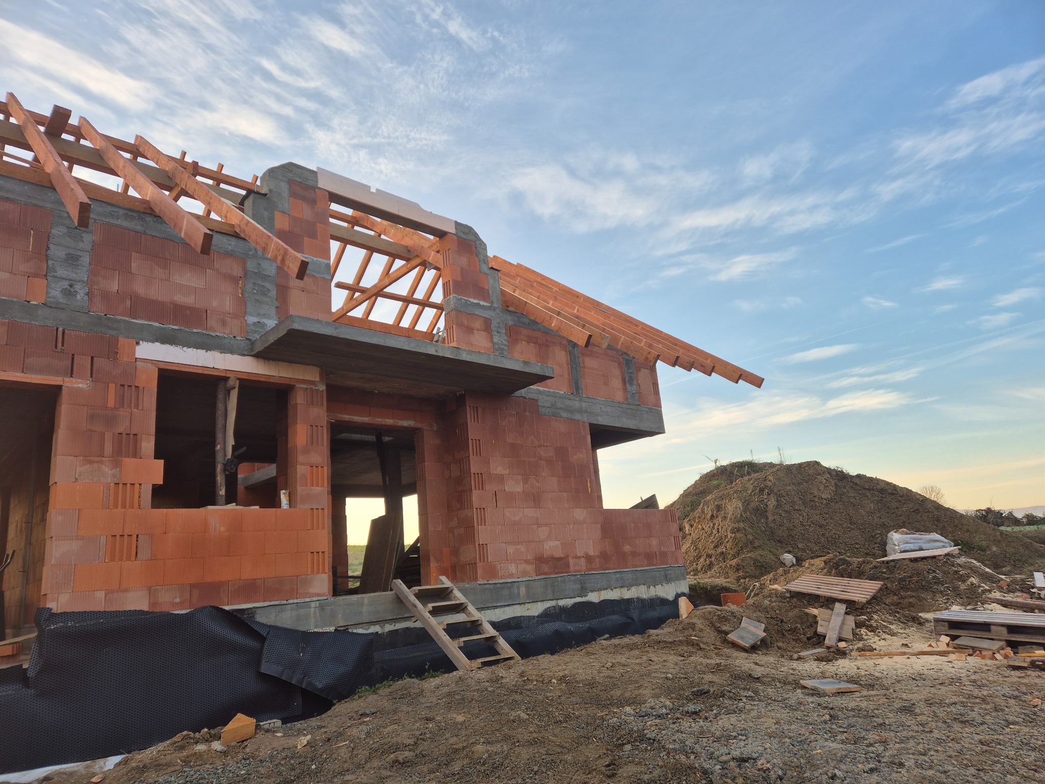 Wznoszenie ścian z czerwonej ceramiki poryzowanej na fundamencie zabezpieczonym folią kubełkową, konstrukcja więźby dachowej w trakcie montażu, drewniana drabina oparta o ścianę, w tle hałda ziemi...