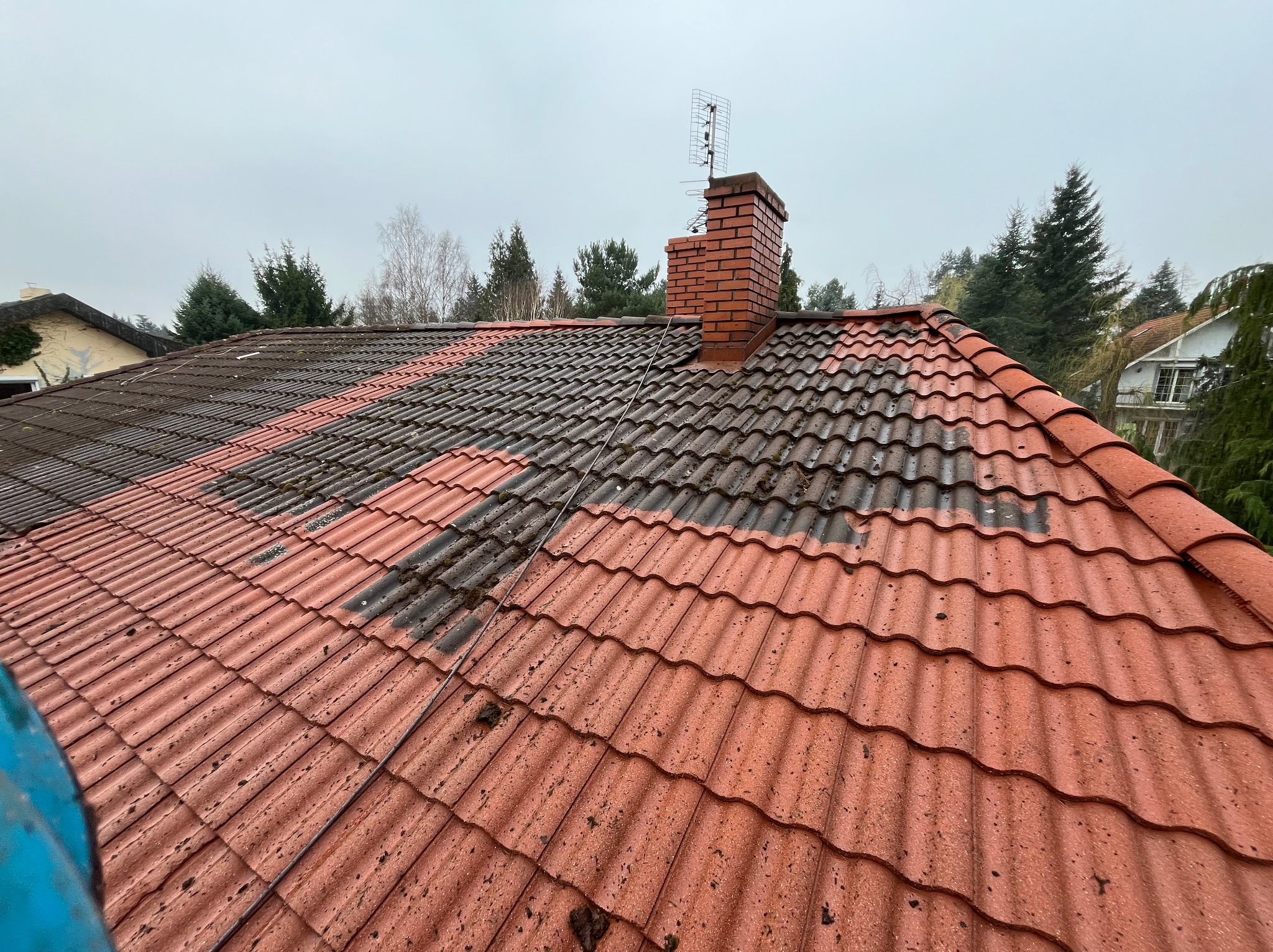 Częściowo oczyszczony dach z czerwonej dachówki ceramicznej, widoczna różnica między czystą a zabrudzoną powierzchnią, ceglany komin z anteną na szczycie.