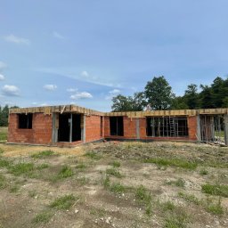 SC-BUD SZYMON ĆWIERZ - Niski budynek w trakcie budowy, wzniesiony z czerwonej cegły, z widocznymi otworami okiennymi i drewnianym szalunkiem stropu, na zewnątrz porośnięty trawą i otoczony drzewami.