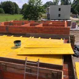 SC-BUD SZYMON ĆWIERZ - Wznoszenie ścian parteru domu z czerwonej cegły ceramicznej, widoczne żółte szalunki stropu i drabina oparta o mur. W tle niezamieszkały budynek w stanie surowym i zielone pole.
