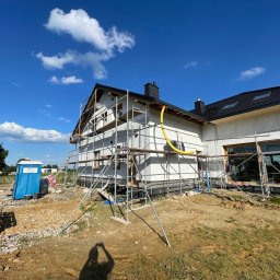 SC-BUD SZYMON ĆWIERZ - Nowoczesny dom w trakcie ocieplania elewacji, z rusztowaniami i widocznym fragmentem terenu budowy. Słoneczny dzień, błękitne niebo z chmurami.