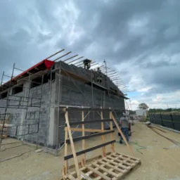Budowa domu z bloczków betonowych: murowane ściany, rusztowania, widoczne krokwie dachu pod czerwonym zabezpieczeniem. Pracownicy na górze, pochmurne niebo.