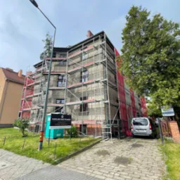 Budynek w rusztowaniach, zabezpieczony siatką, prawdopodobnie w trakcie renowacji elewacji. Na pierwszym planie przenośna toaleta i reklama kancelarii prawnej.