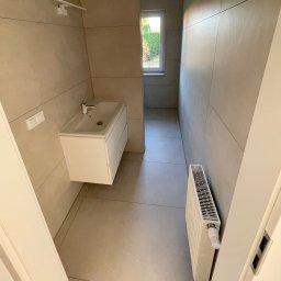 SC-BUD SZYMON ĆWIERZ - Jasna łazienka po remoncie: beżowe płytki na ścianach i podłodze, biała szafka z umywalką, grzejnik. Widok przez otwarte drzwi i okno na zieleń.