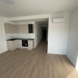 SC-BUD SZYMON ĆWIERZ - Jasna, minimalistyczna kuchnia z beżowymi szafkami i czarnym blatem, widok na korytarz. Klimatyzator na ścianie. Podłoga z paneli imitujących drewno.