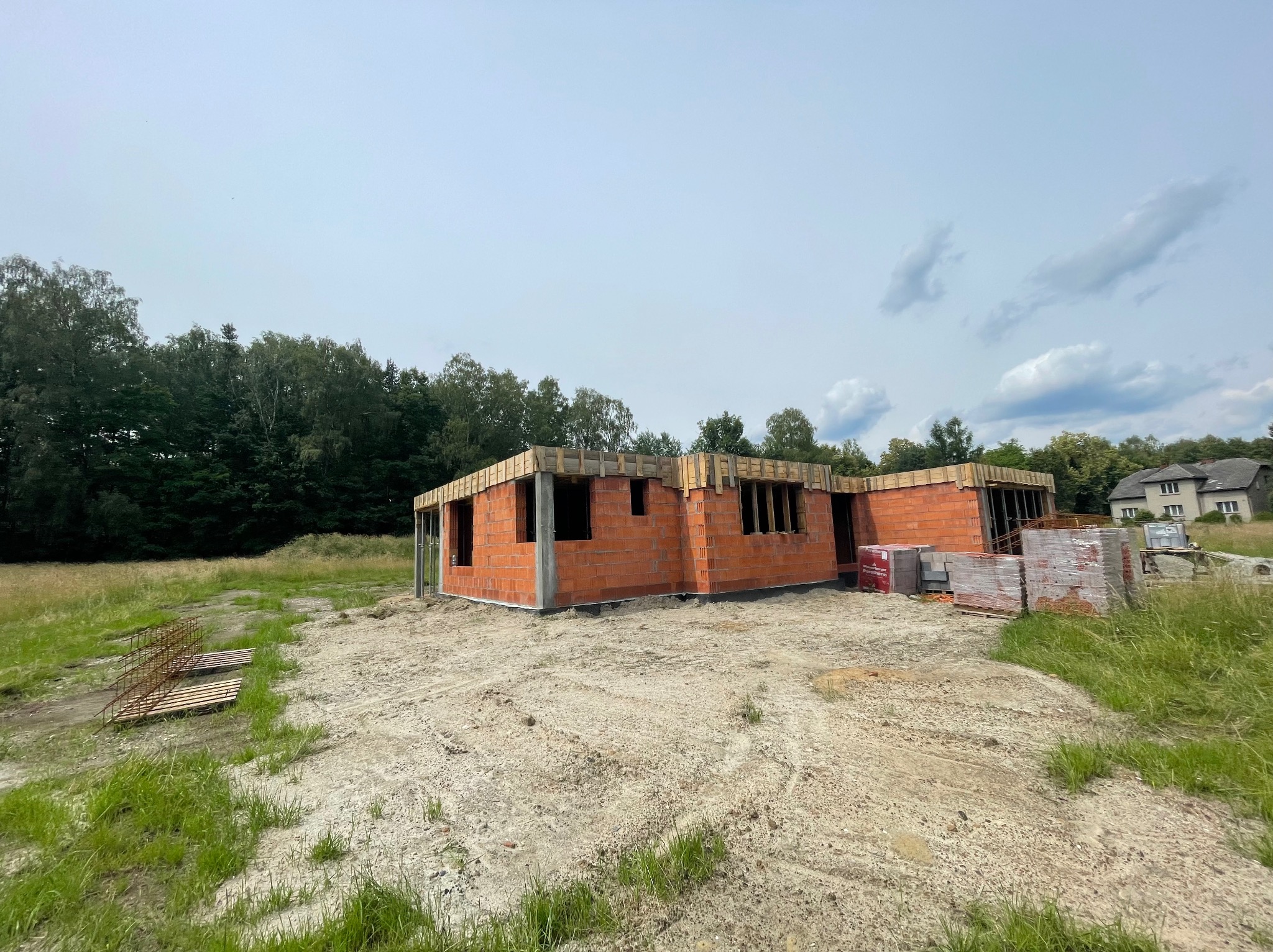 Surowy stan domu murowanego z czerwonej cegły, widoczne drewniane nadproża okienne, palety z cegłami obok, otoczenie: trawa i drzewa.