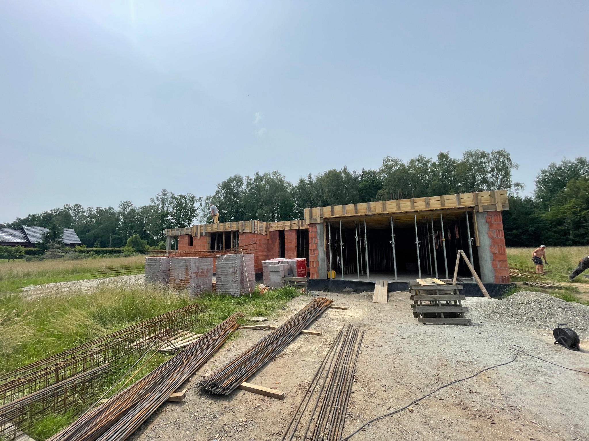 Budowa domu w stanie surowym otwartym: murowane ściany z czerwonej cegły, drewniane szalunki stropu, zbrojenia na ziemi, widoczni pracownicy na tle zieleni i nieba.