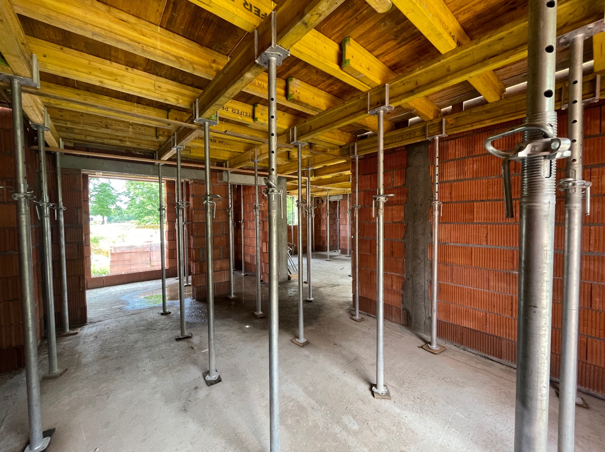 Wnętrze budowanego domu z cegły, z widocznymi podporami stropowymi z żółtymi deskami szalunkowymi, surowy beton na podłodze.