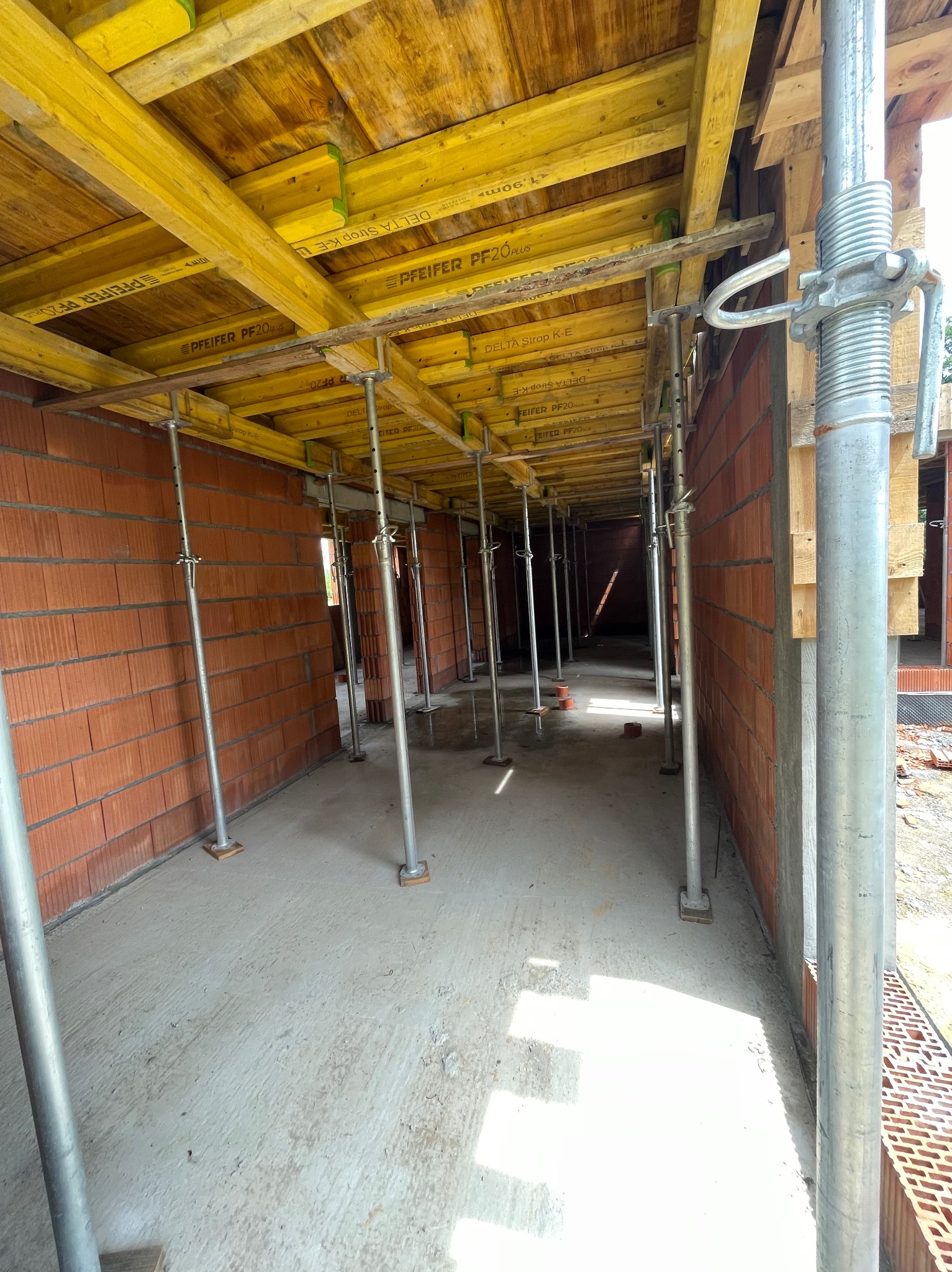 Wnętrze w trakcie budowy z widocznymi ścianami z cegły, żółtym szalunkiem stropowym wspartym na metalowych podporach i betonową posadzką.