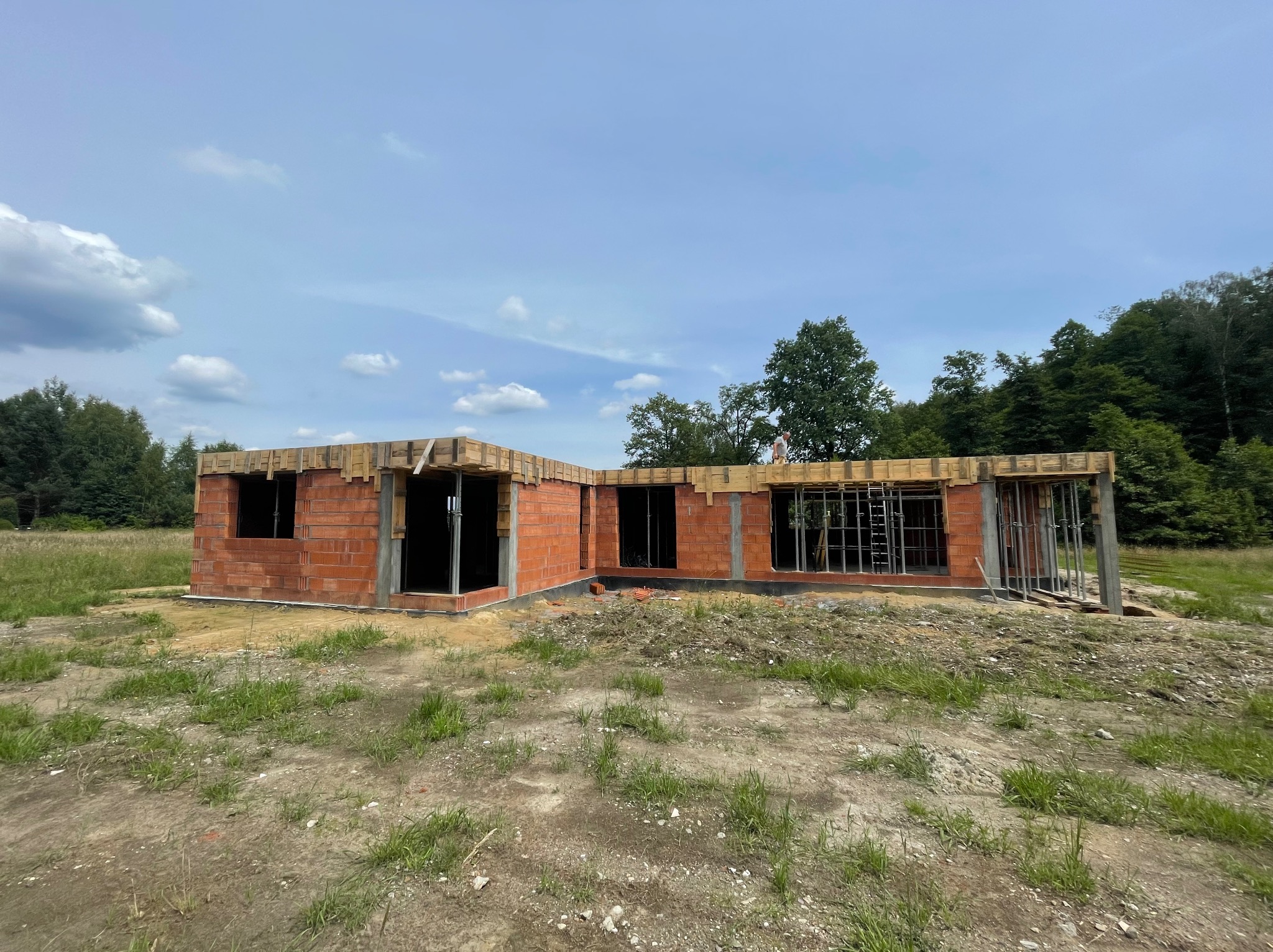 Niski budynek w trakcie budowy, wzniesiony z czerwonej cegły, z widocznymi otworami okiennymi i drewnianym szalunkiem stropu, na zewnątrz porośnięty trawą i otoczony drzewami.