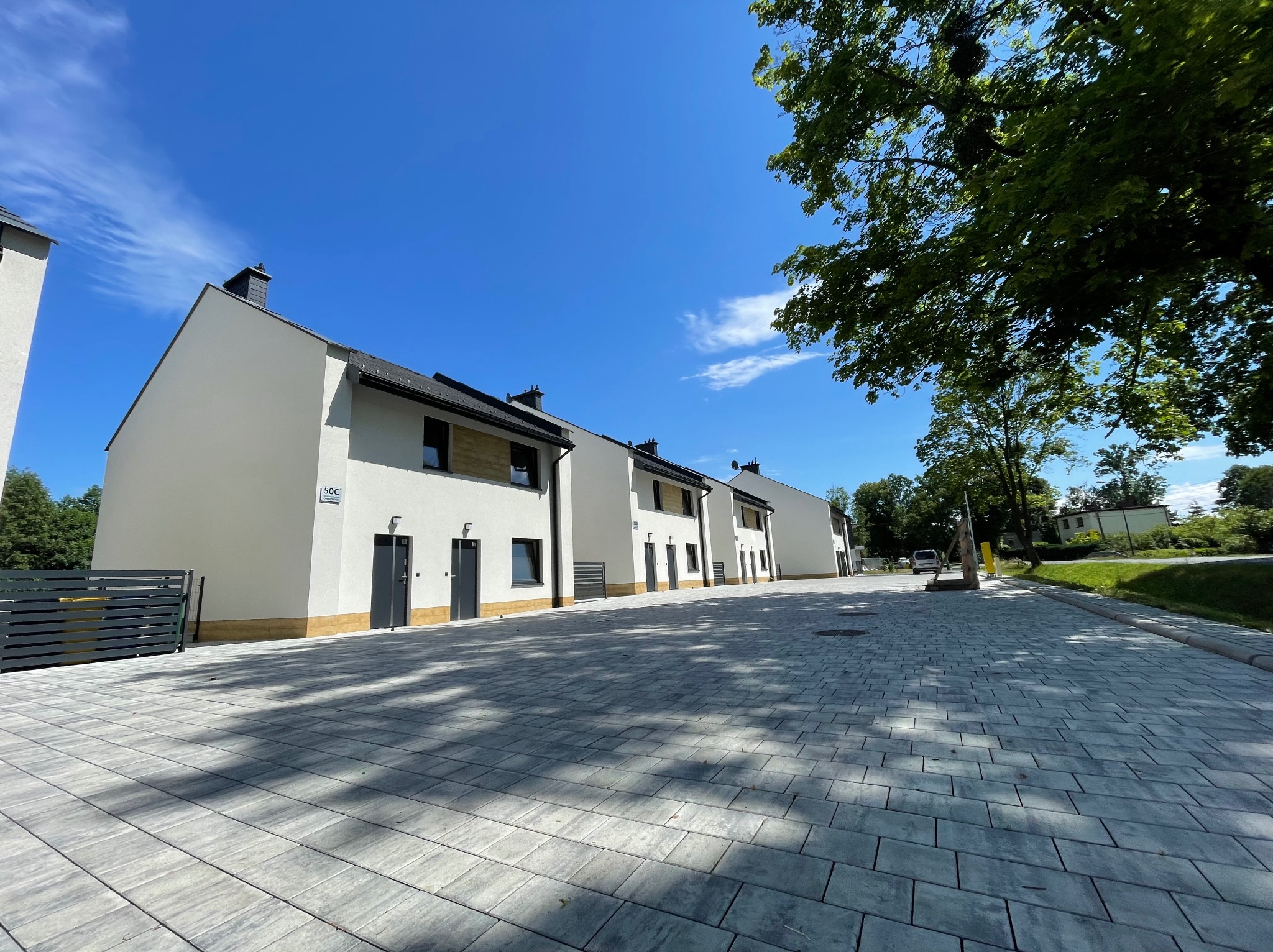 Szereg nowo wybudowanych, nowoczesnych domów jednorodzinnych w zabudowie szeregowej z białymi elewacjami, ciemnymi dachami i szarą kostką brukową na podjeździe, sfotografowanych w słoneczny dzień...