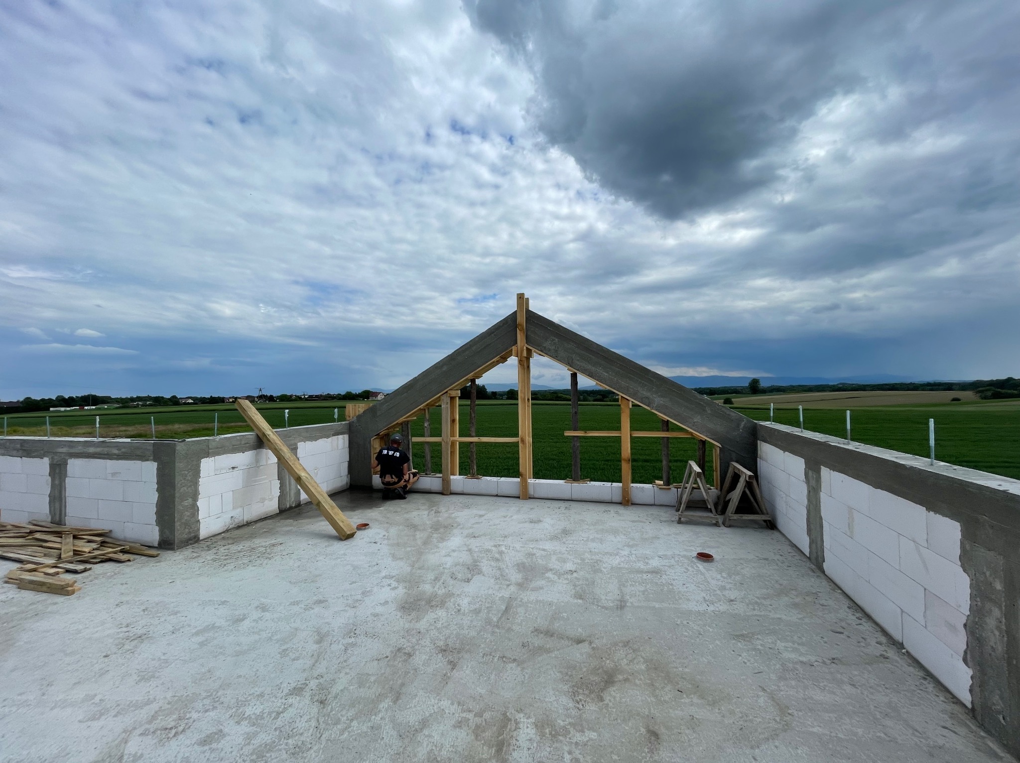 Wznoszenie konstrukcji dachu na surowym murze domu z betonu komórkowego, widok na zielone pola i pochmurne niebo w tle.