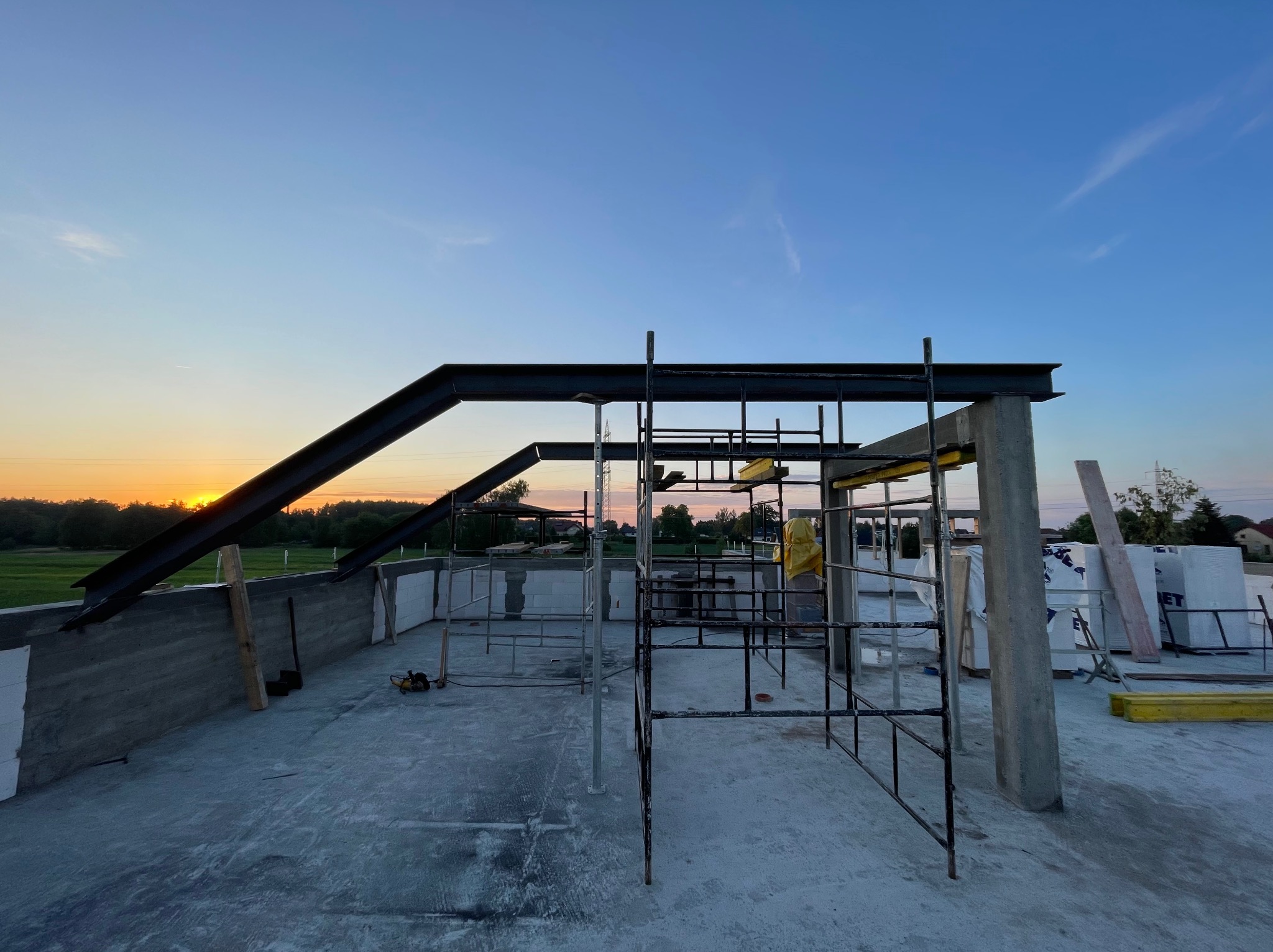 Konstrukcja stalowa z elementami rusztowania na surowym stropie budynku w trakcie budowy, widok na zachodzące słońce i krajobraz za budynkiem.