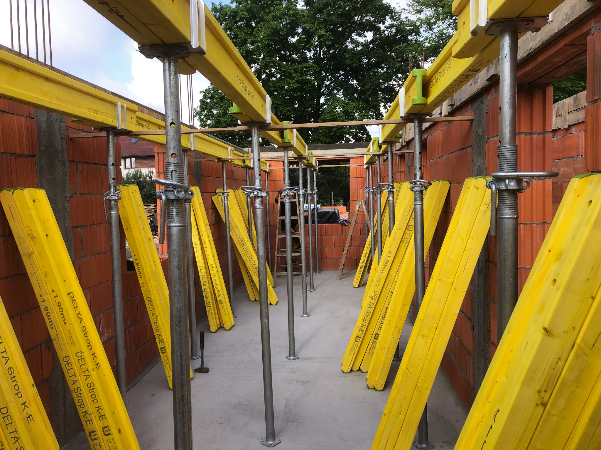 Wnętrze budynku w trakcie budowy z widocznymi ścianami z czerwonej cegły, stalowymi podporami i żółtymi belkami stropowymi DELTA Strop K-E, na betonowej posadzce leży młotek.