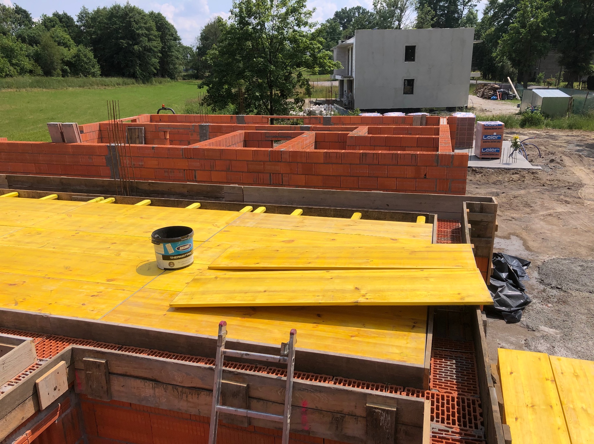 Wznoszenie ścian parteru domu z czerwonej cegły ceramicznej, widoczne żółte szalunki stropu i drabina oparta o mur. W tle niezamieszkały budynek w stanie surowym i zielone pole.