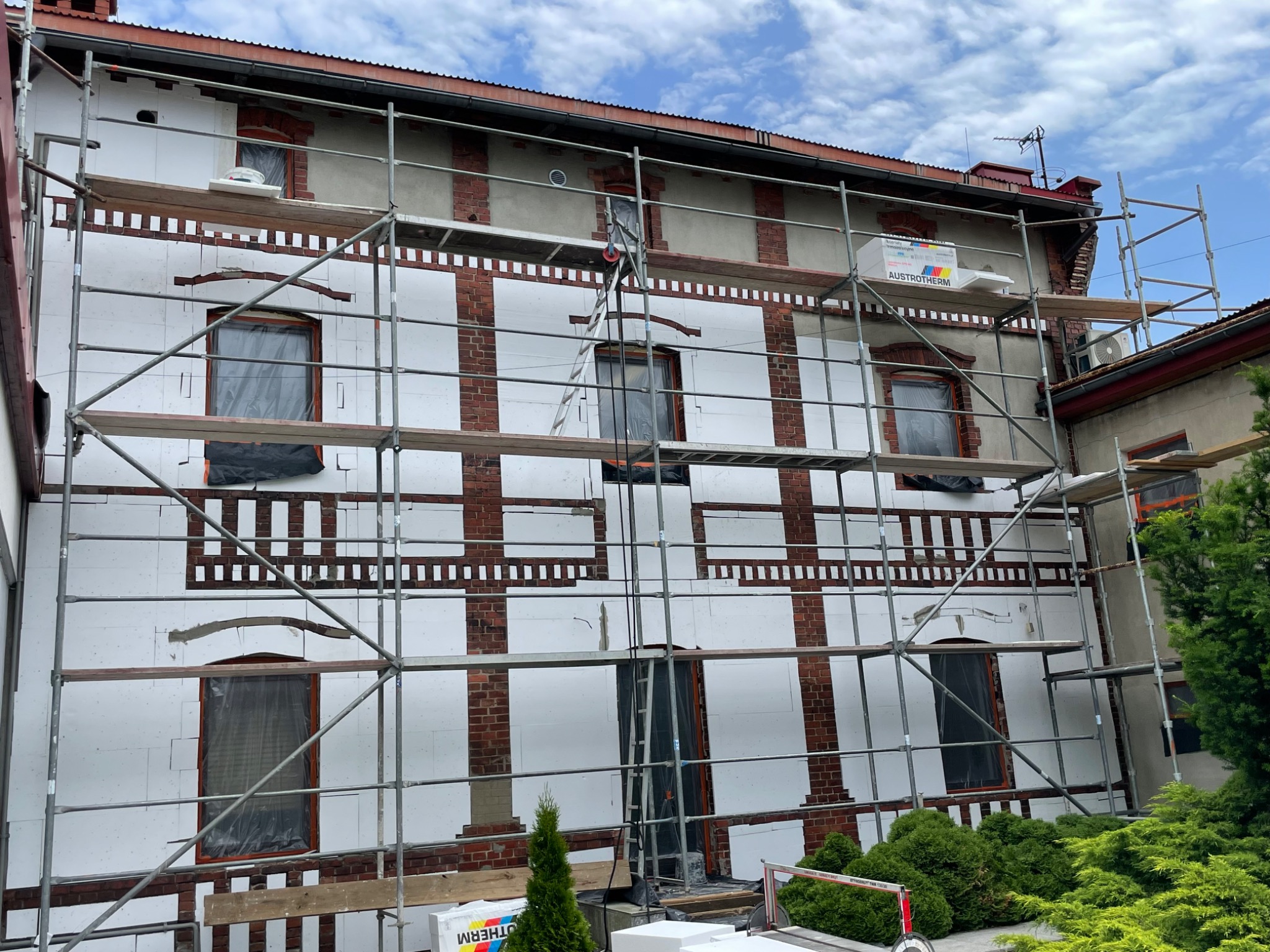 Elewacja budynku w trakcie ocieplania styropianem, widoczne rusztowania i okna zabezpieczone folią. Częściowo ocieplona ściana z odsłoniętą starą cegłą.