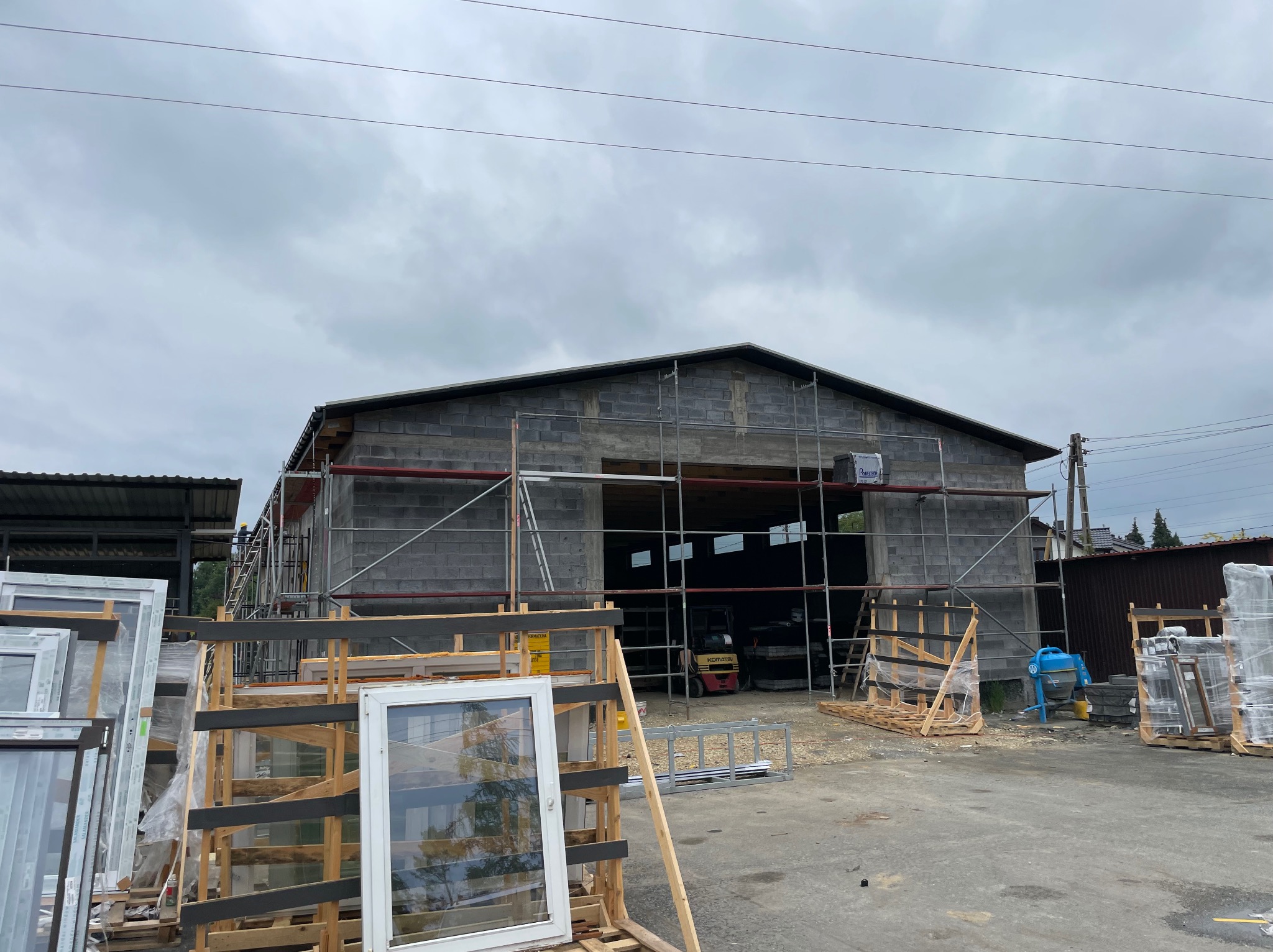Budynek w trakcie murowania z szarej cegły, otoczony rusztowaniami. Na pierwszym planie okna w drewnianych ramach, a w tle widać wózek widłowy.
