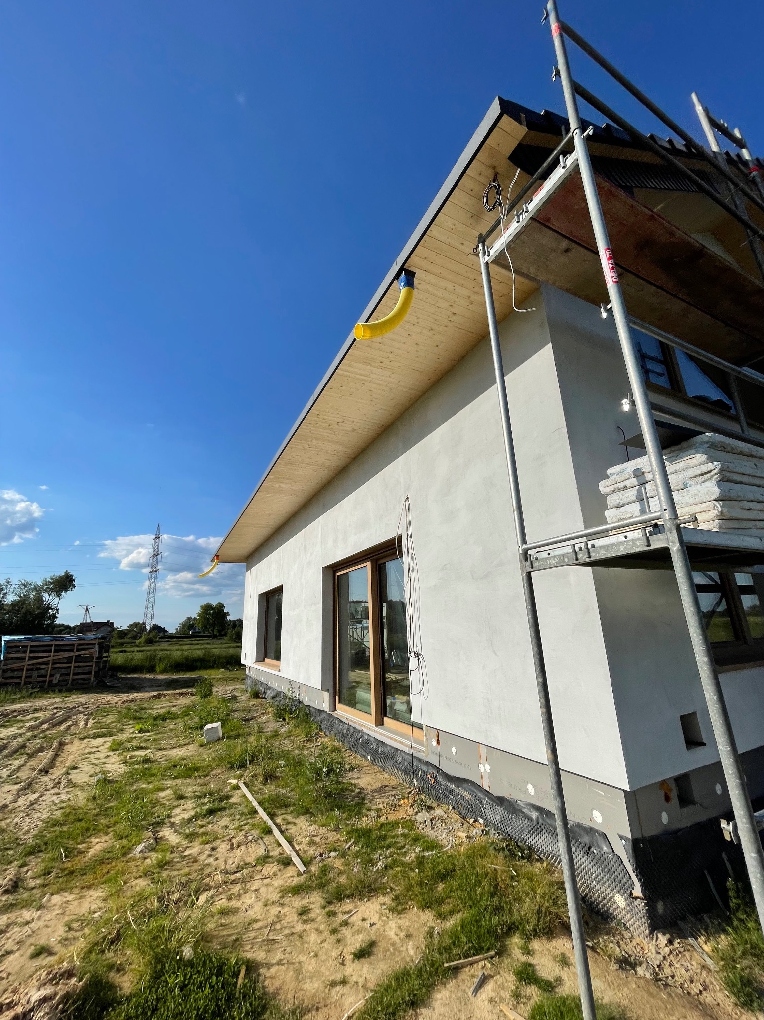 Dom jednorodzinny w trakcie ocieplania, widoczne rusztowanie, okna z drewnianą stolarką i ocieplenie. Na dachu żółta rura wentylacyjna. Błękitne niebo w tle.
