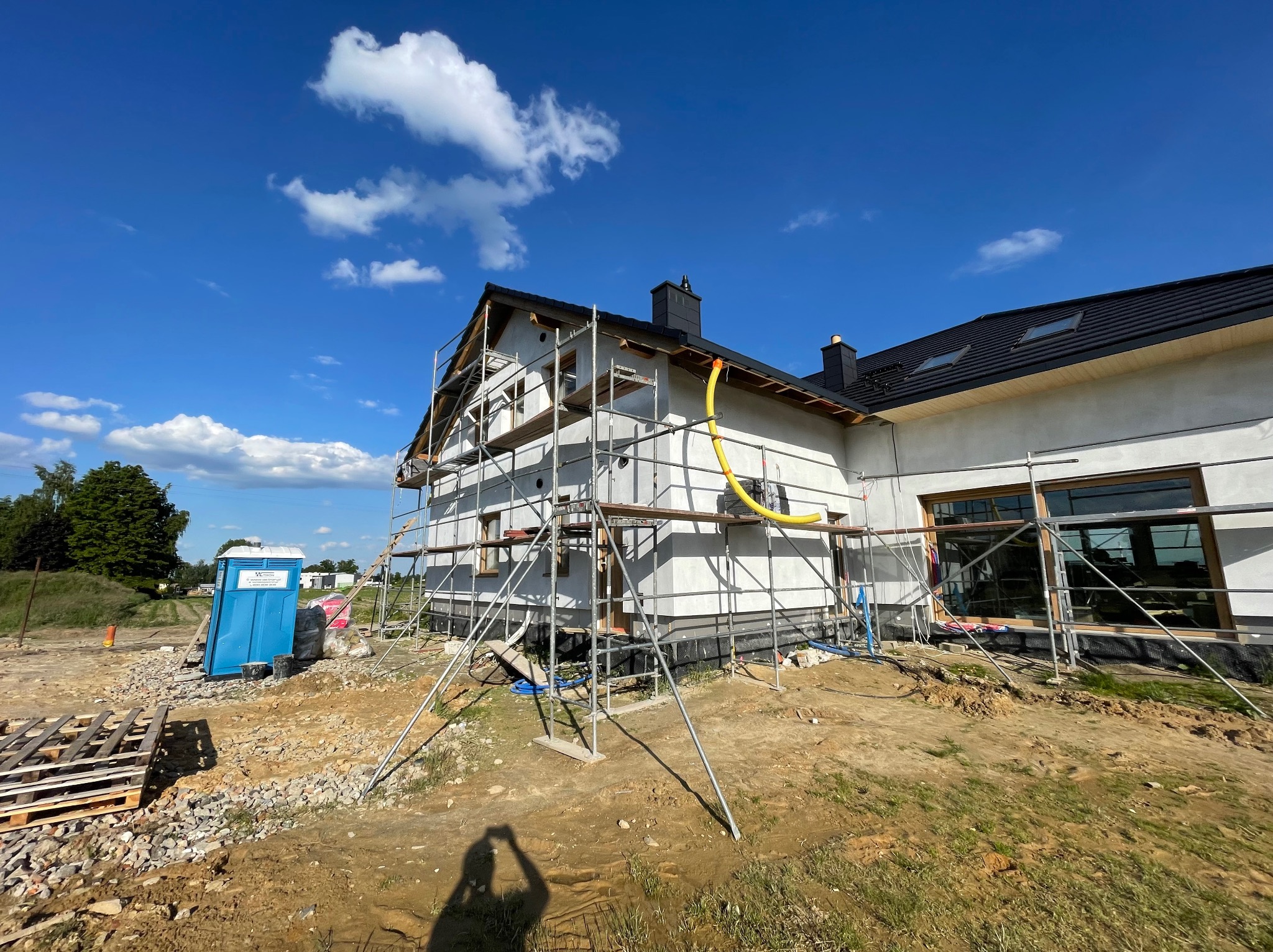 Nowoczesny dom w trakcie ocieplania elewacji, z rusztowaniami i widocznym fragmentem terenu budowy. Słoneczny dzień, błękitne niebo z chmurami.
