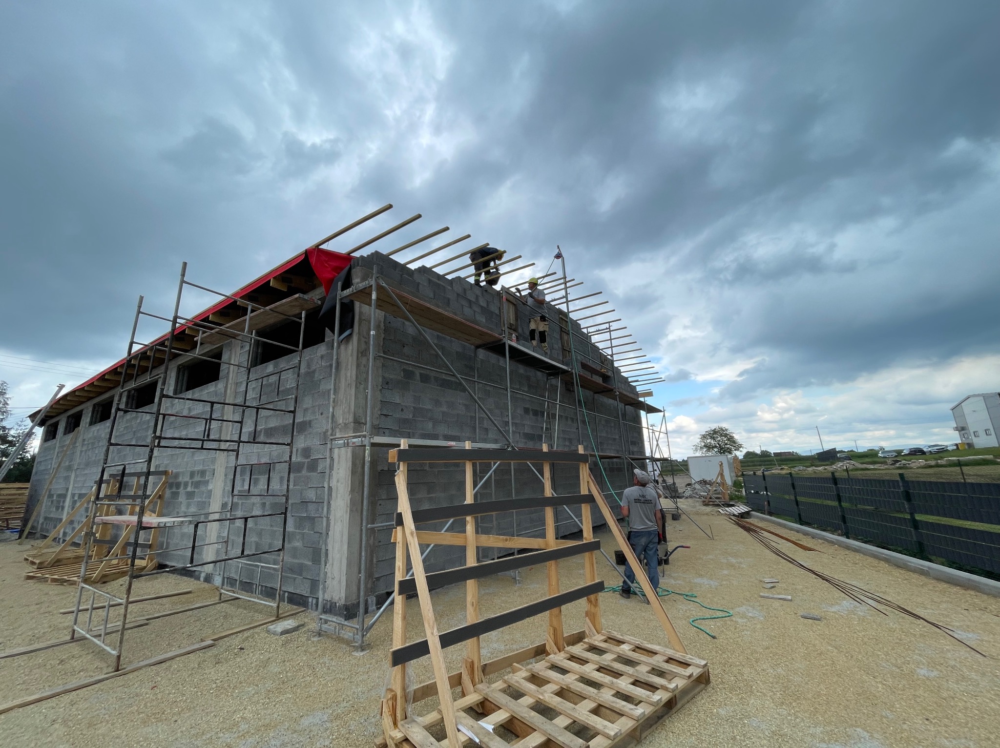 Budowa domu z bloczków betonowych: murowane ściany, rusztowania, widoczne krokwie dachu pod czerwonym zabezpieczeniem. Pracownicy na górze, pochmurne niebo.