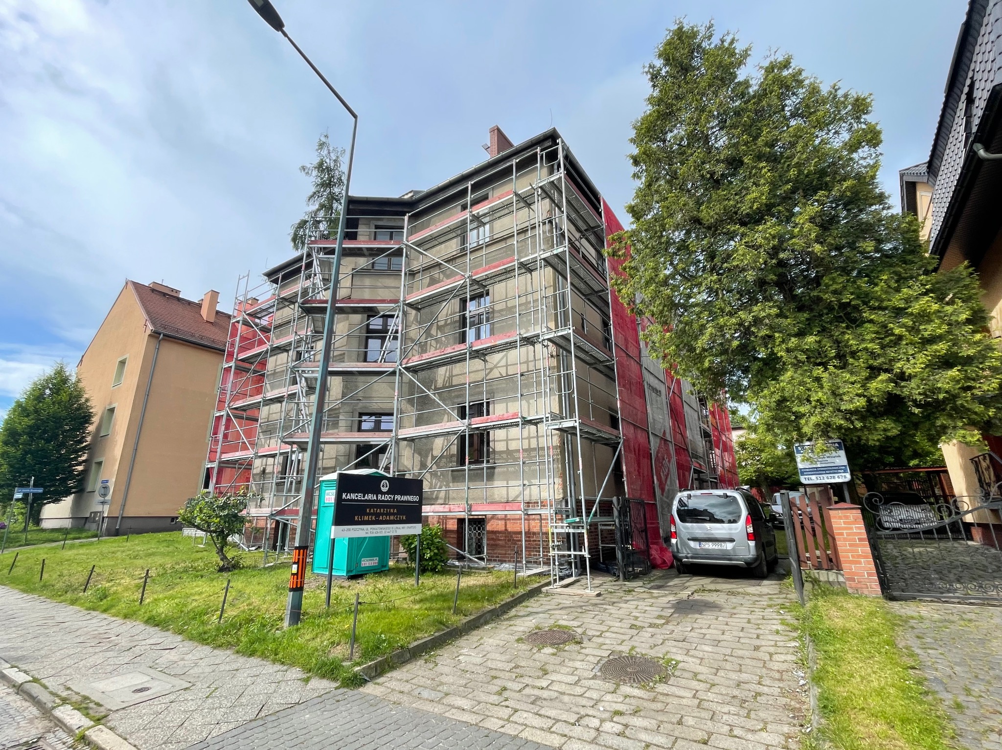 Budynek w rusztowaniach, zabezpieczony siatką, prawdopodobnie w trakcie renowacji elewacji. Na pierwszym planie przenośna toaleta i reklama kancelarii prawnej.