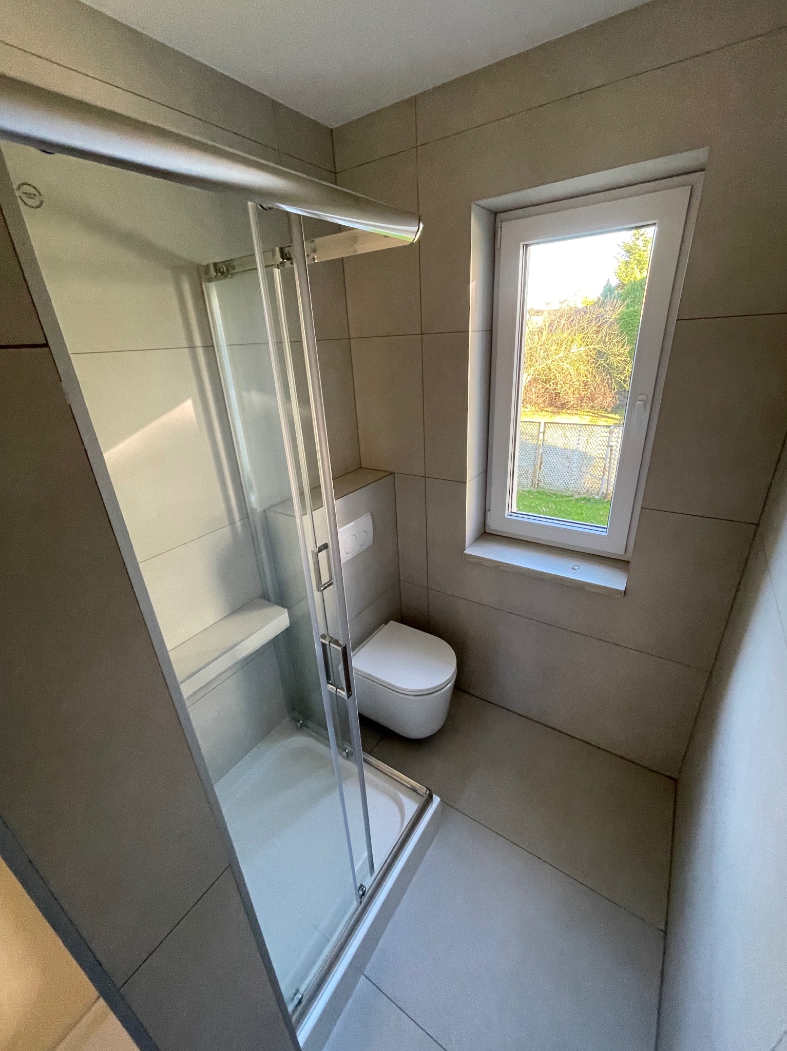 Nowoczesna łazienka w szarościach: kabina prysznicowa ze szklanymi drzwiami, podwieszana toaleta i okno z widokiem na zieleń. Minimalistyczny design, płytki wielkoformatowe.