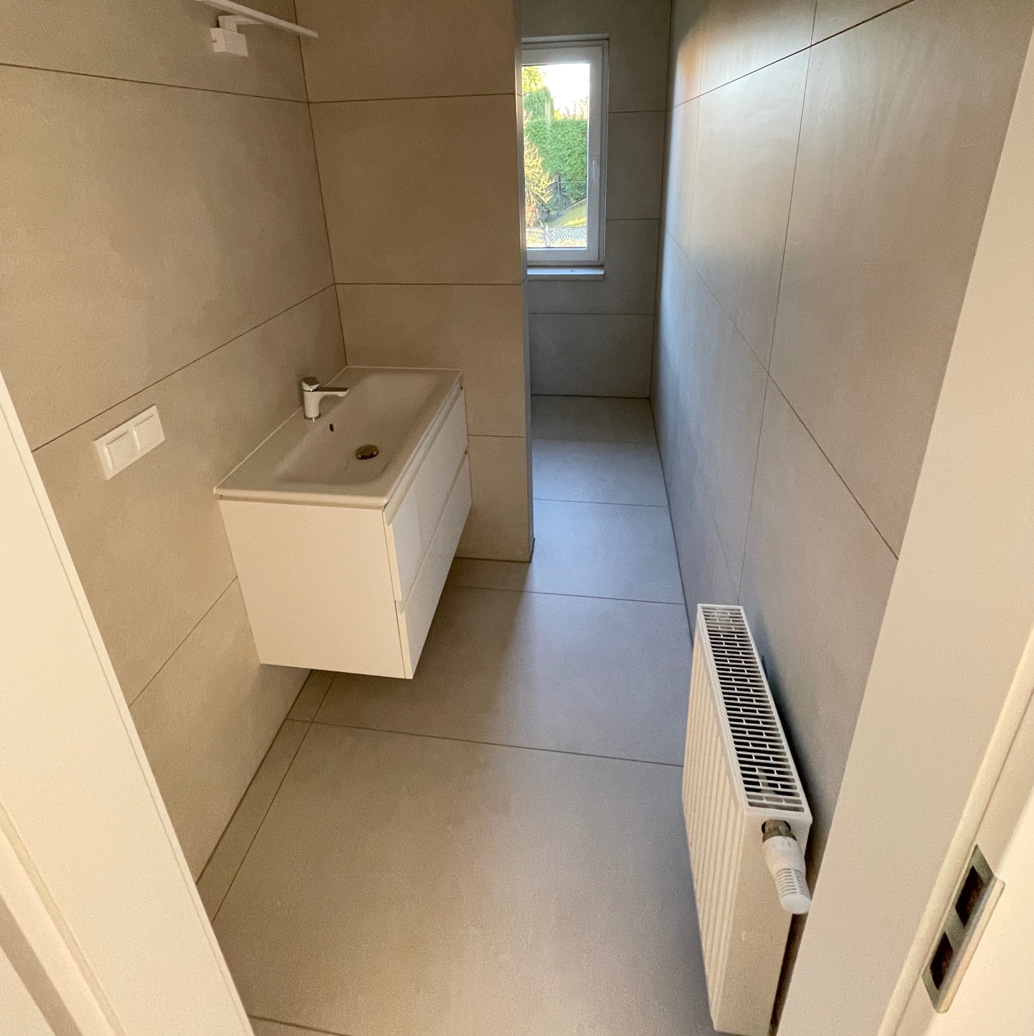 Jasna łazienka po remoncie: beżowe płytki na ścianach i podłodze, biała szafka z umywalką, grzejnik. Widok przez otwarte drzwi i okno na zieleń.